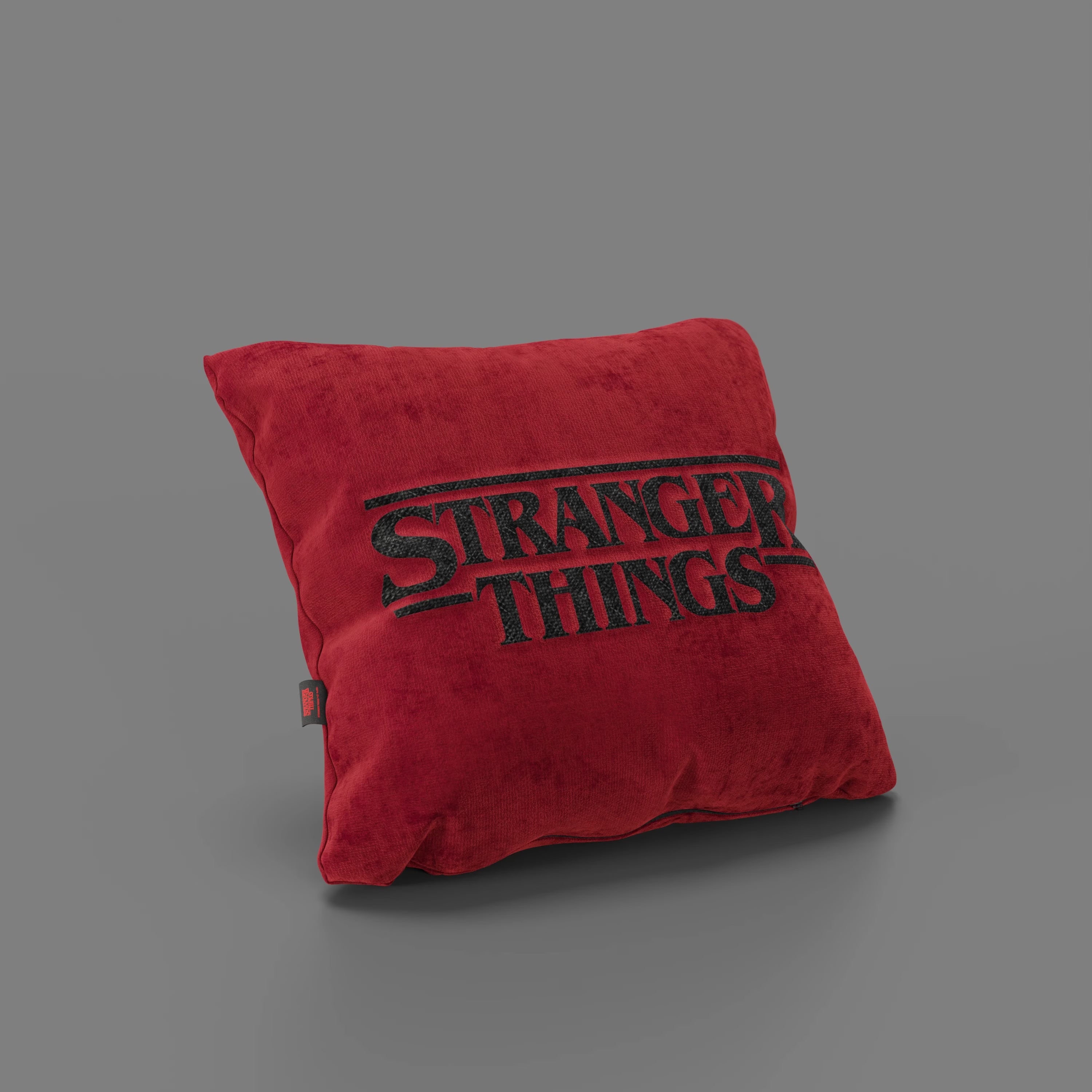 Il cuscino Cuscino Stranger Things Limited Edition 45x45 di Divani.Store, di forma quadrata e di colore rosso, poggia su una superficie grigia e presenta il logo "Stranger Things" ricamato e una piccola etichetta laterale nera. Realizzato in tessuto antimacchia.