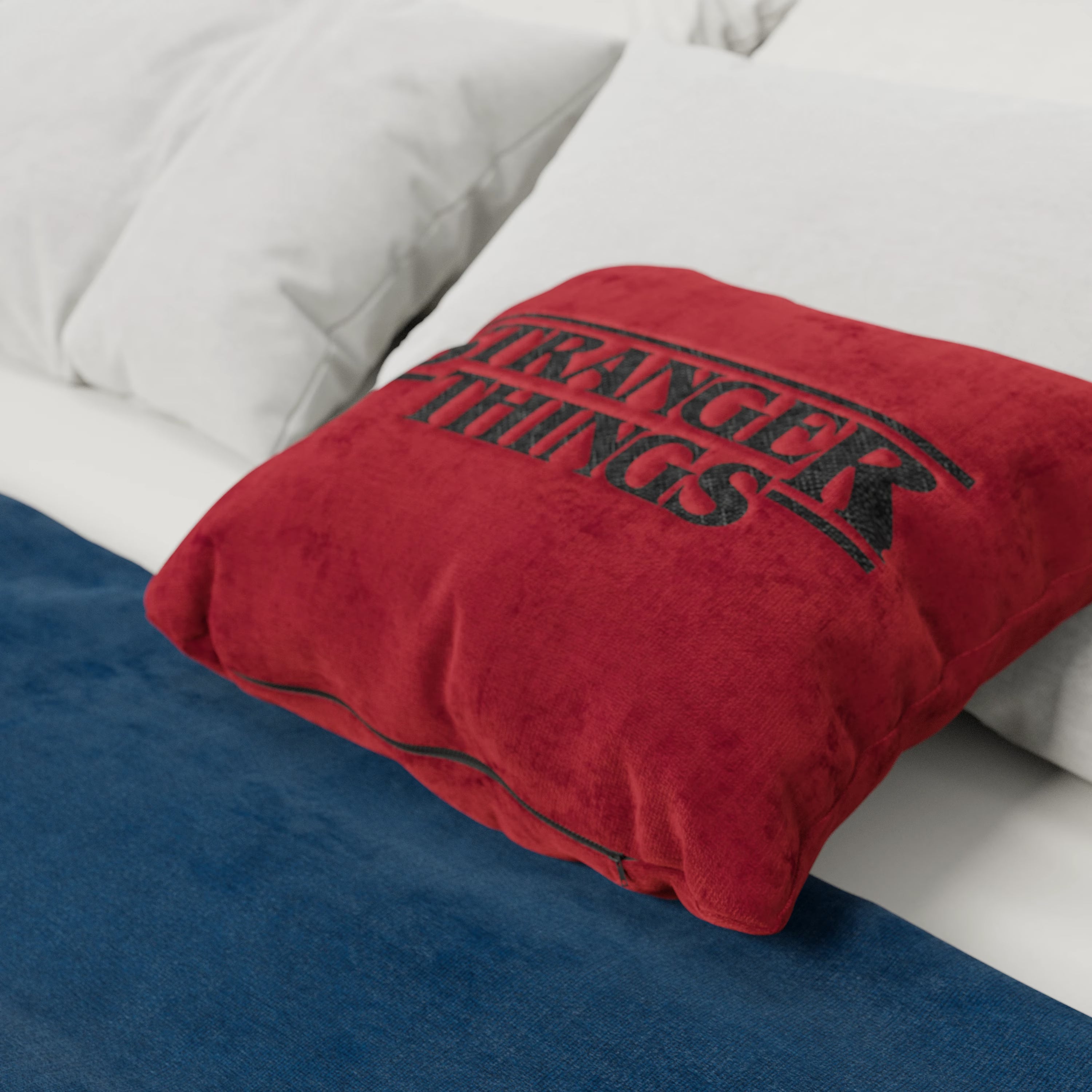 Un Cuscino Stranger Things Limited Edition 45x45 di Divani.Store in rosso, con logo ricamato e tessuto antimacchia, aggiunge un tocco accogliente e ispirato a Netflix a un letto con cuscini bianchi e una coperta blu scuro.