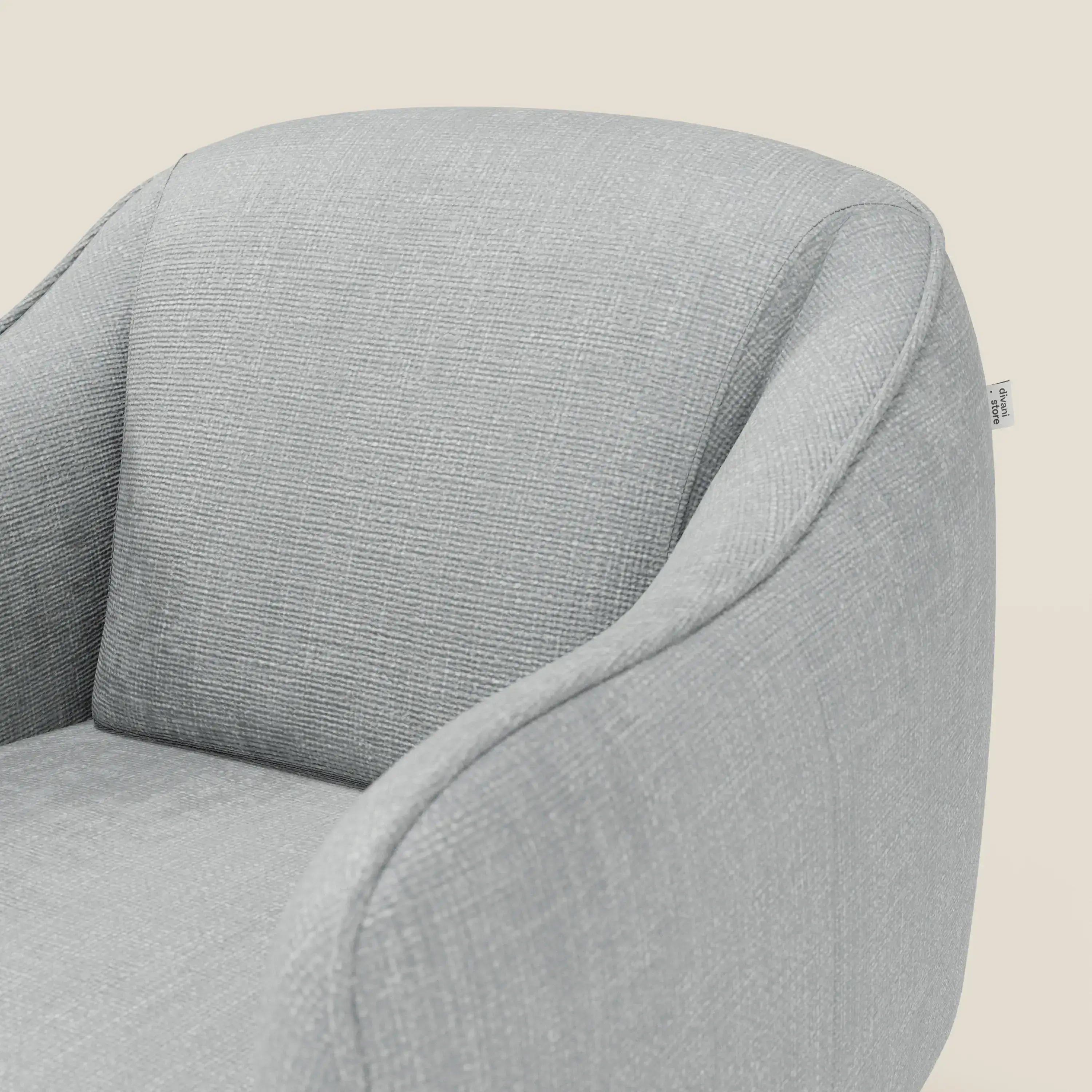 Primo piano di Avela Poltroncina di Divani.Store, con schienale curvo e braccioli in morbido tessuto T06 ghiaccio, resistente all'acqua, la cui trama e la piccola etichetta laterale evidenziano il comfort senza tempo su uno sfondo bianco sporco.