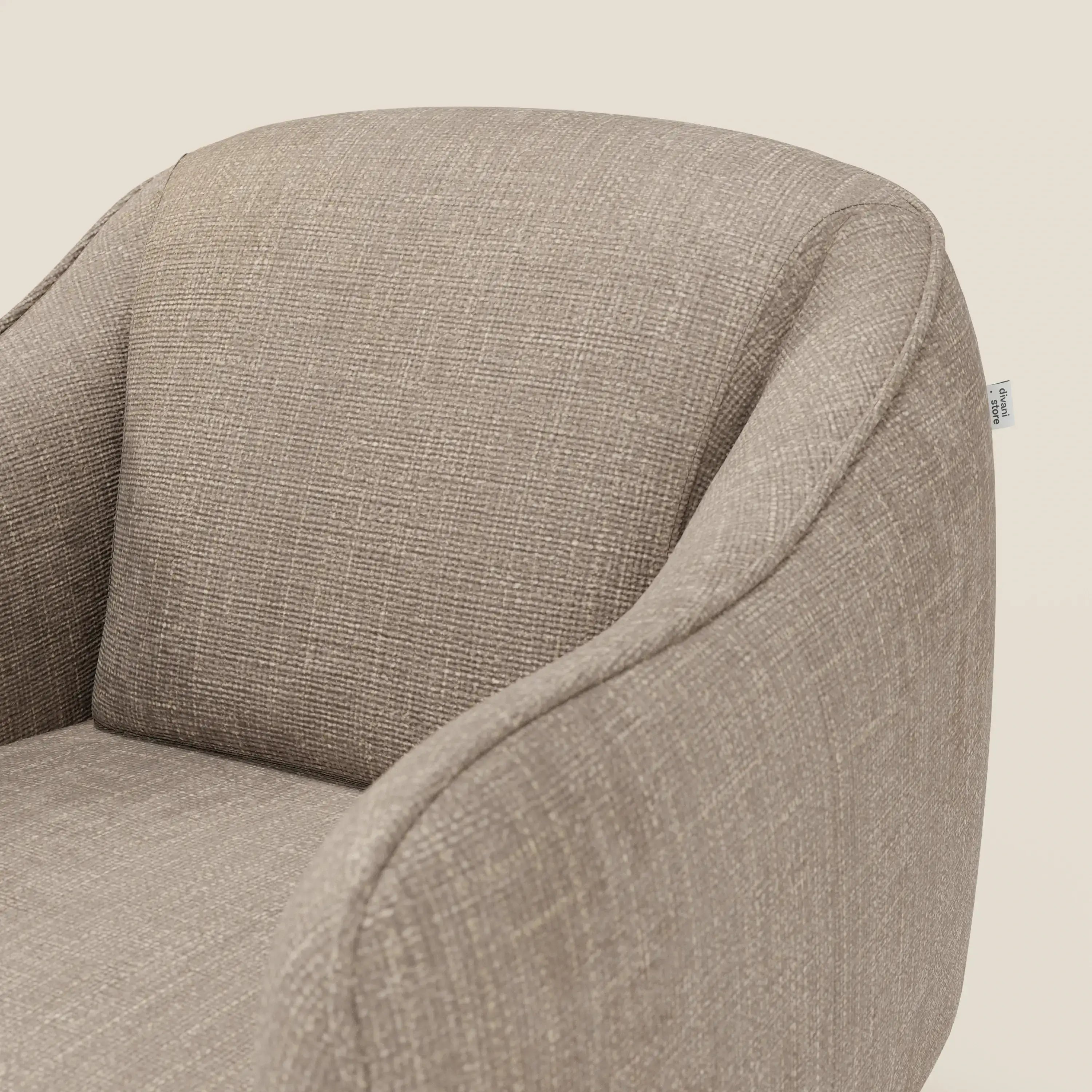 Un primo piano di Avela Poltroncina di Divani.Store in morbido tessuto tortora T06 idrorepellente mette in evidenza lo schienale curvo, i braccioli arrotondati e le raffinate cuciture su uno sfondo crema chiaro.
