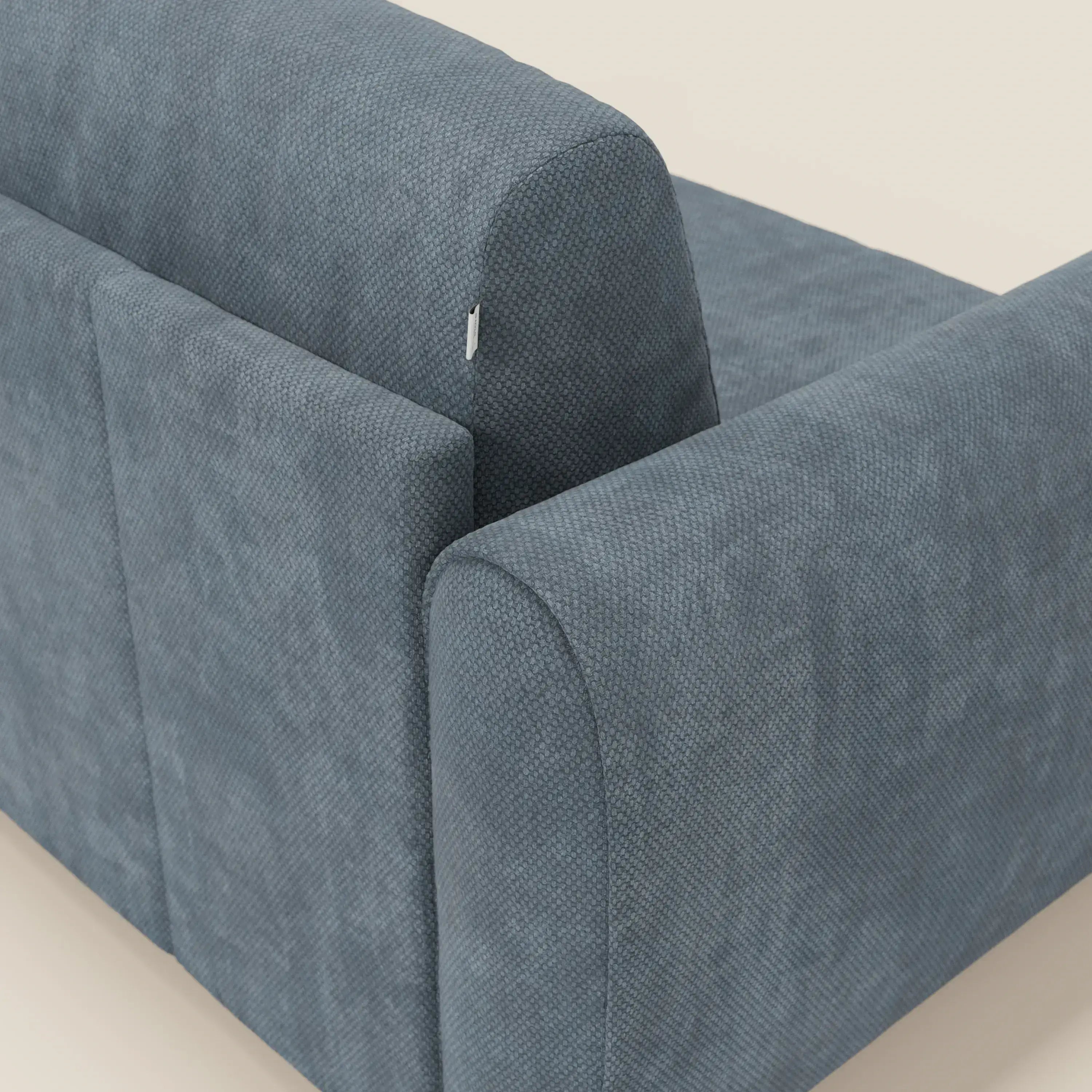 Un primo piano del divano letto moderno Kaleo di Divani.Store in tessuto impermeabile T26 carta da zucchero mostra il suo tessuto blu testurizzato e un'etichetta di manutenzione bianca vicino alla cucitura dello schienale, sullo sfondo di un semplice bianco sporco.