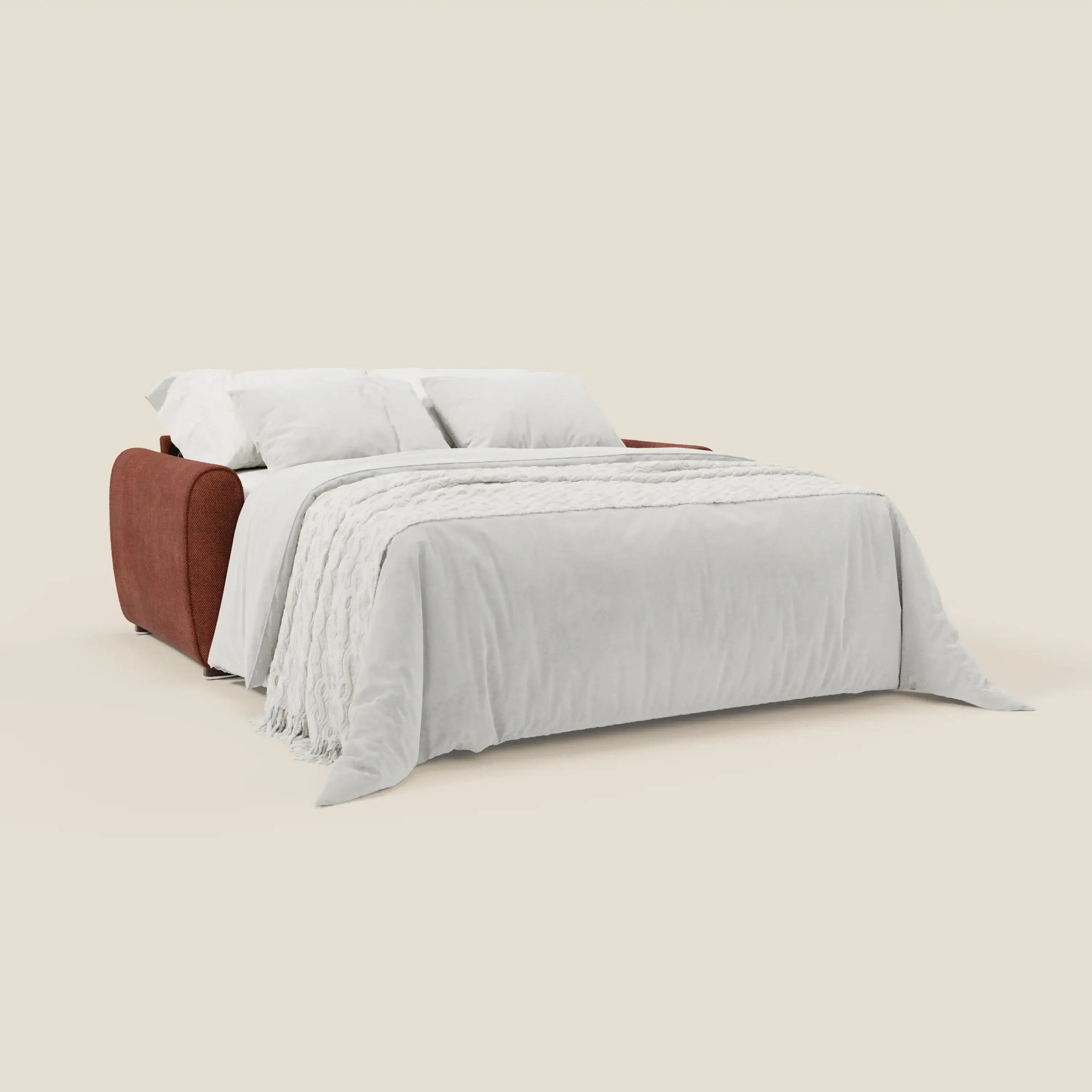 Il divano letto moderno Kaleo di Divani.Store, in elegante tessuto impermeabile T26 arancione ruggine, è aperto a letto con lenzuolo bianco, due cuscini bianchi e una coperta bianca con frange su sfondo beige chiaro.