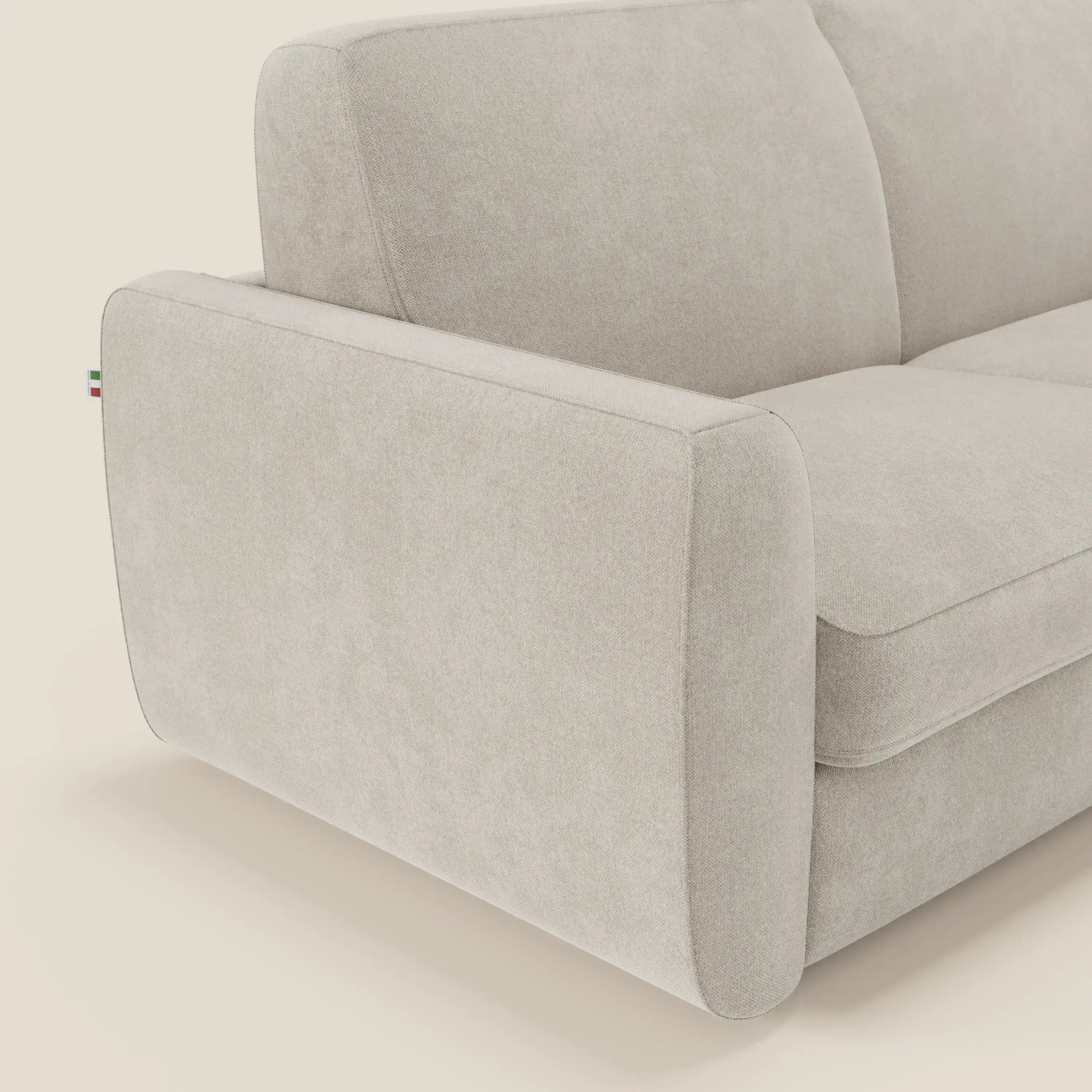 Un primo piano del divano letto sfoderabile Leonida di Divani.Store in tessuto smacchiabile T23 panna mostra i cuscini in tessuto, il bracciolo sinistro e la texture morbida, con una targhetta tricolore che sottolinea il meccanismo acciaio rinforzato.