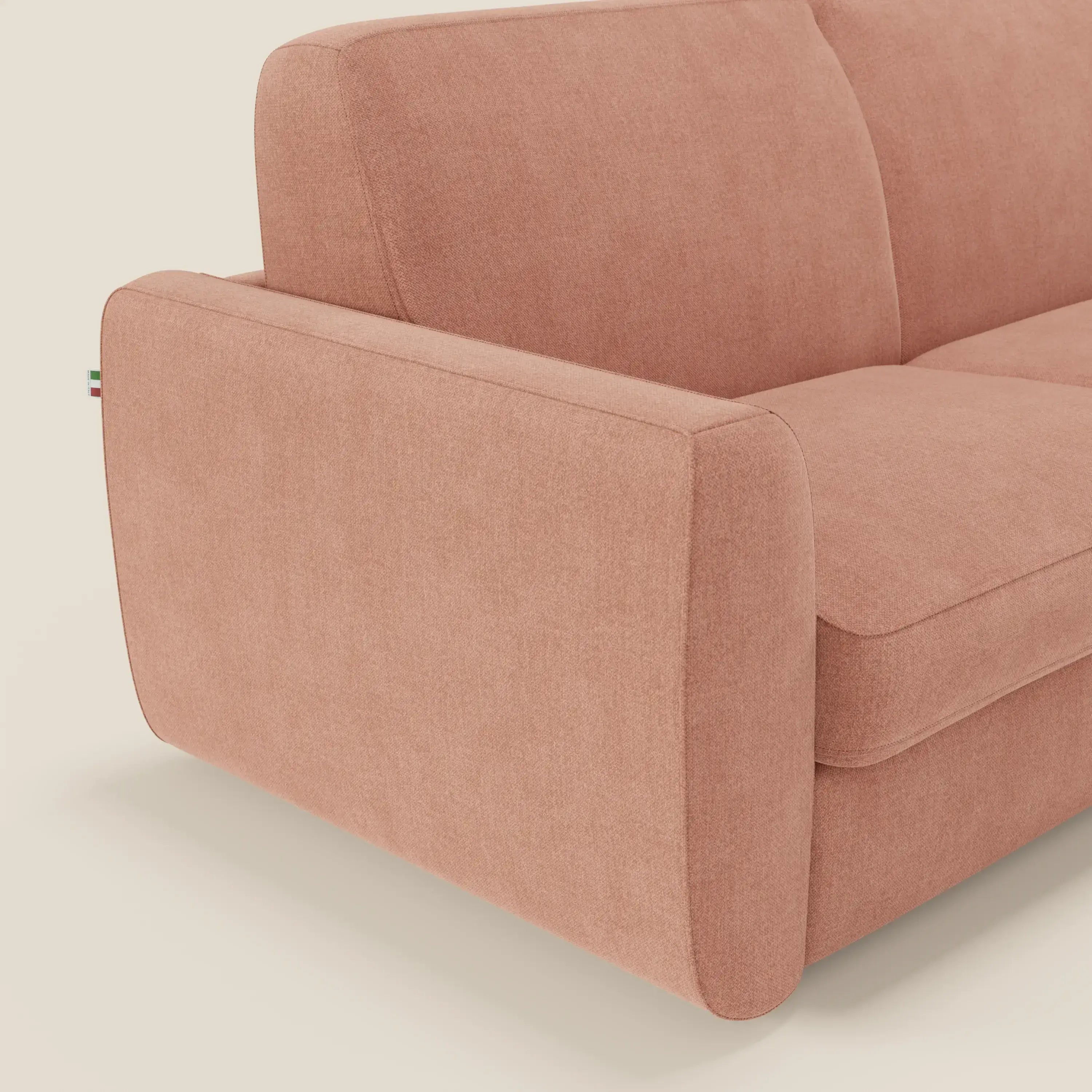 Primo piano del bracciolo sinistro e dei cuscini del divano letto sfoderabile Leonida di Divani.Store, rivestito in tessuto Smacchiabile T23 pesca, con etichetta tricolore e meccanismo acciaio rinforzato per una maggiore durata.