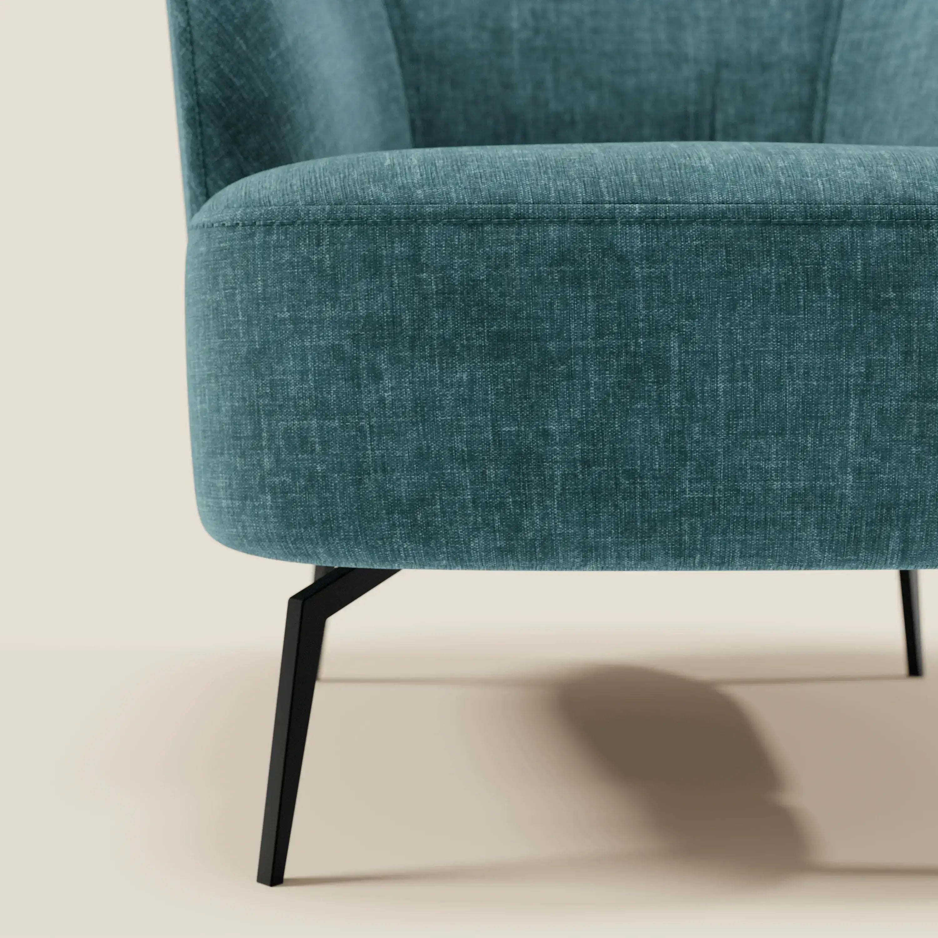 Primo piano di Renoa Poltroncina di Divani.Store, rivestita in tessuto smacchiabile T24 petrolio, con schienale e seduta curvi per il massimo comfort. Le sottili gambe angolate in metallo nero contrastano elegantemente con il fondo beige chiaro.