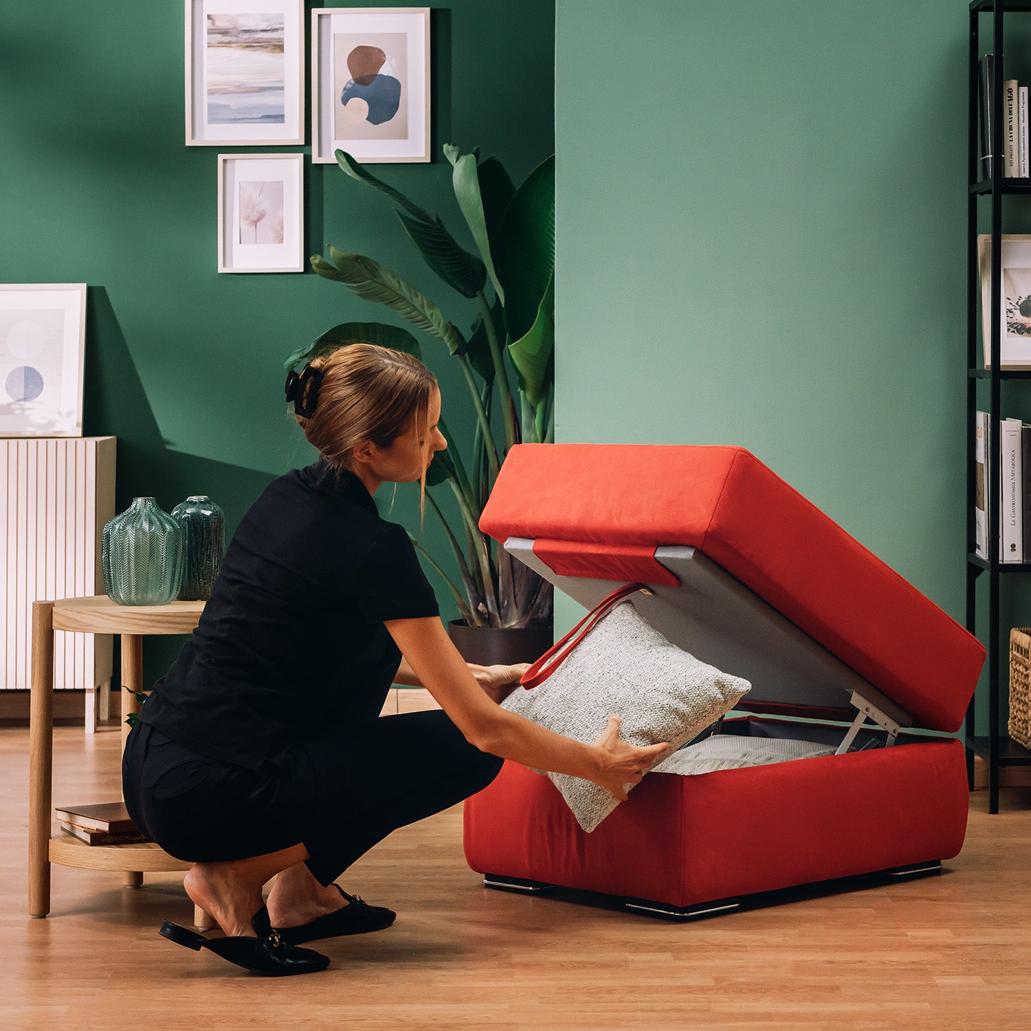 Una donna vestita di nero si inginocchia su un pavimento di legno e inserisce un cuscino grigio chiaro nel pouf Sun apribile contenitore in microfibra smacchiabile T11 di Divani.Store, circondato da pareti verdi, opere d'arte incorniciate, piante, un tavolo di legno e decorazioni in vetro.