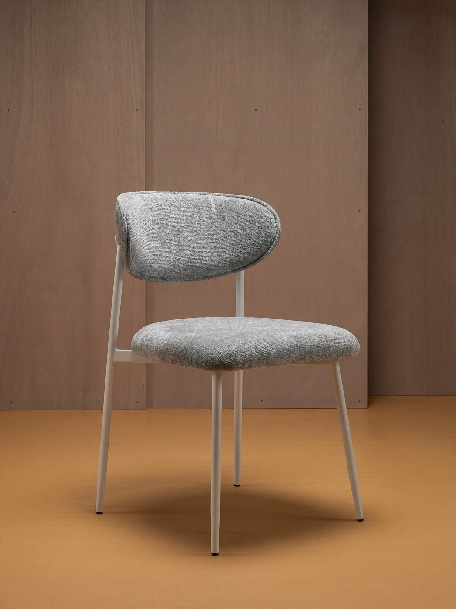 Una sedia moderna con seduta imbottita di colore grigio chiaro e dalla trama ruvida, schienale arrotondato e gambe in metallo bianco, che fa da contrasto con una parete rivestita in legno e un pavimento color senape.