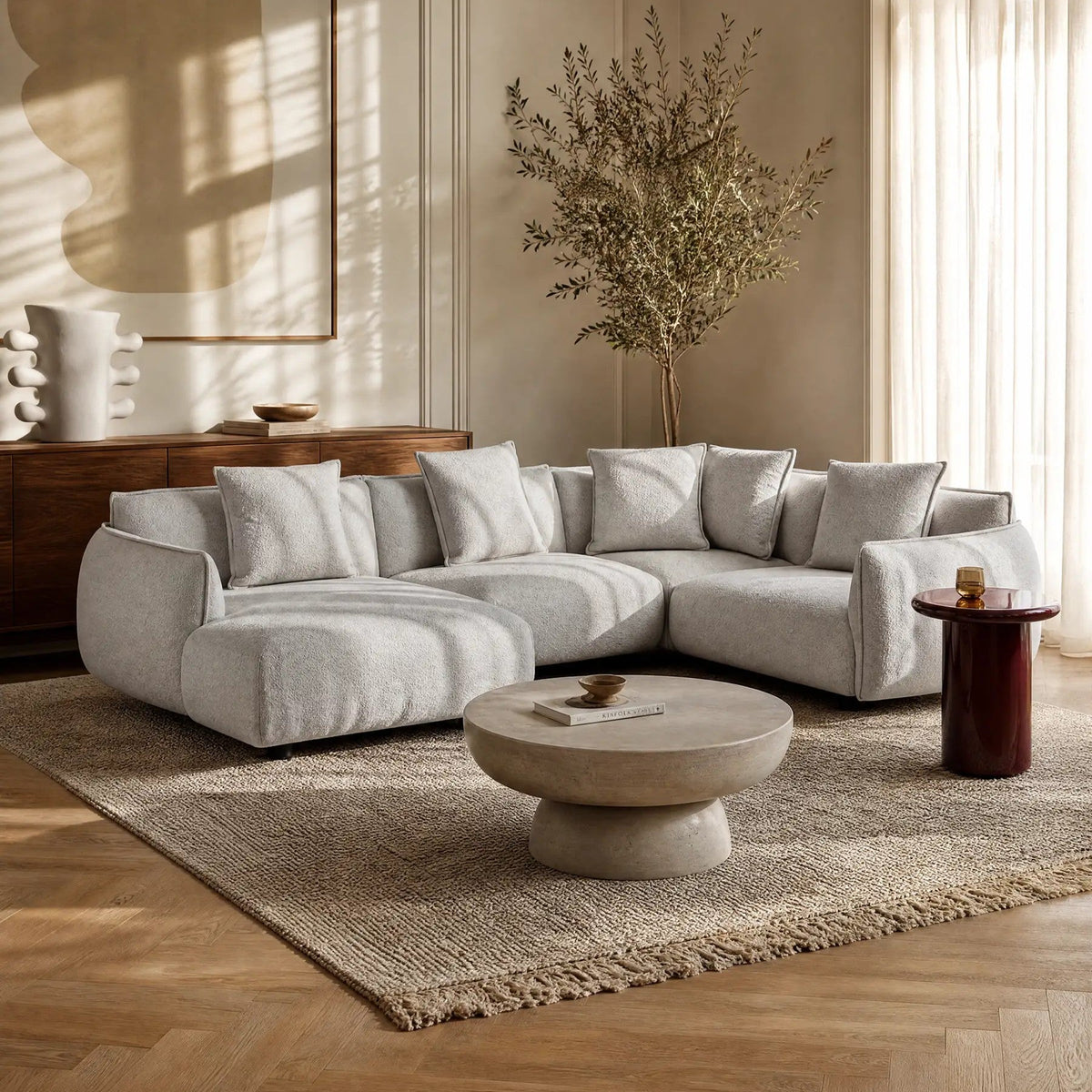 Soggiorno moderno con divano componibile grigio chiaro, tavolino rotondo in pietra, tappeto con trama in rilievo, credenza in legno, arredamento minimalista e luce naturale che entra dalle ampie finestre con tende trasparenti.