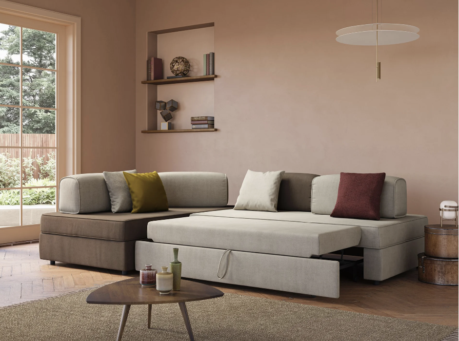 Un soggiorno moderno con un divano componibile beige con contenitore estraibile, quattro cuscini in colori neutri, un tavolino rotondo in legno, mensole a muro con decorazioni, una grande finestra e una lampada a sospensione.