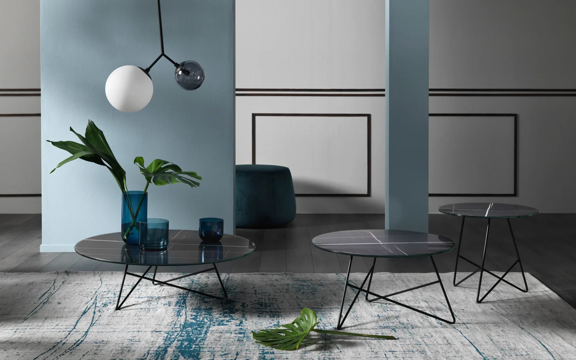 Soggiorno moderno con tre tavoli rotondi neri con sottili gambe in metallo, bicchieri decorativi blu, foglie verdi in un vaso, una foglia caduta su un tappeto fantasia e lampade a sospensione geometriche.