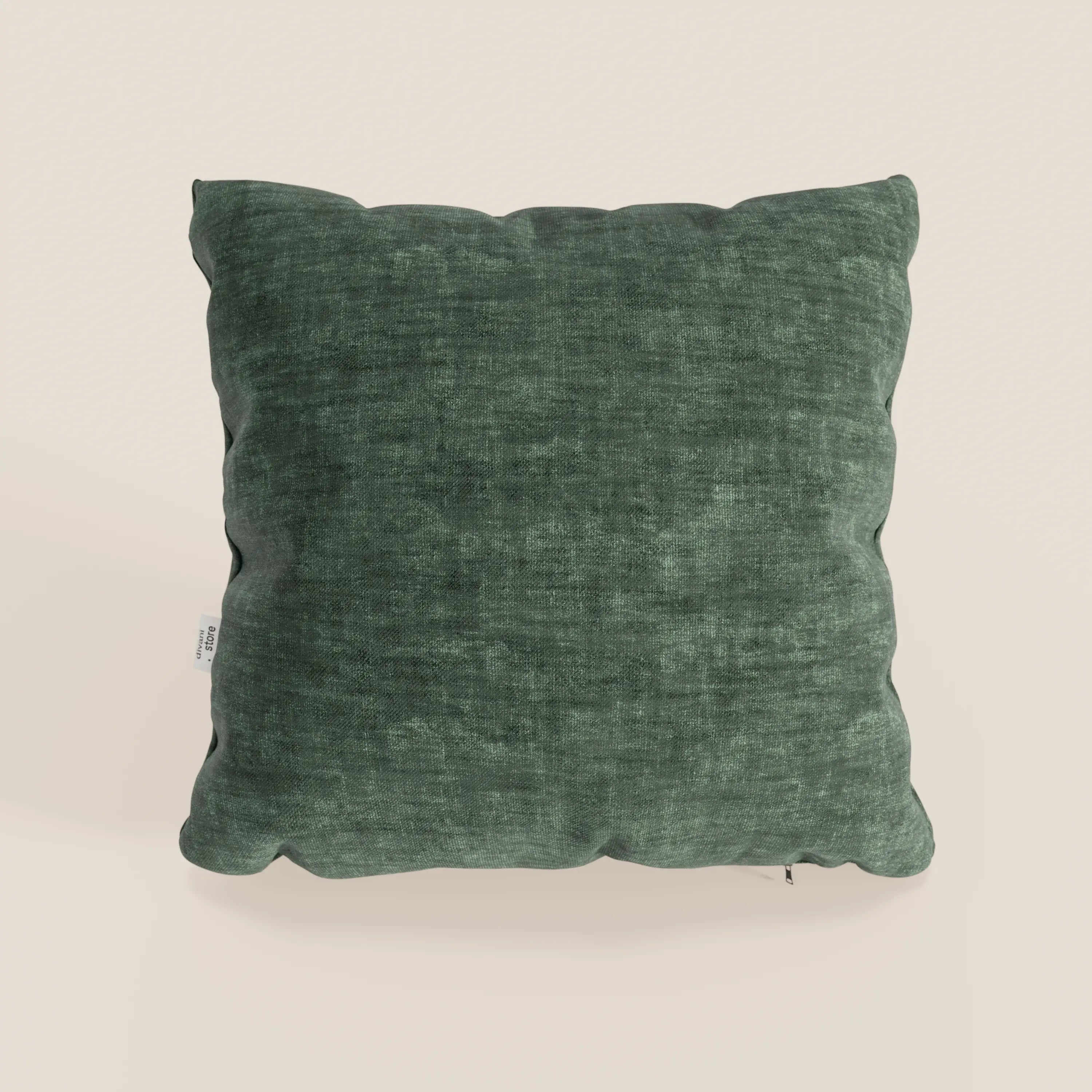 Un cuscino quadrato Sofia verde scuro di Divani.Store, in morbido velluto antimacchia e impermeabile (Easy Clean T35), poggia su una superficie beige chiaro, con un aspetto abbondante e una leggera piega e una piccola etichetta bianca sulla cucitura sinistra.