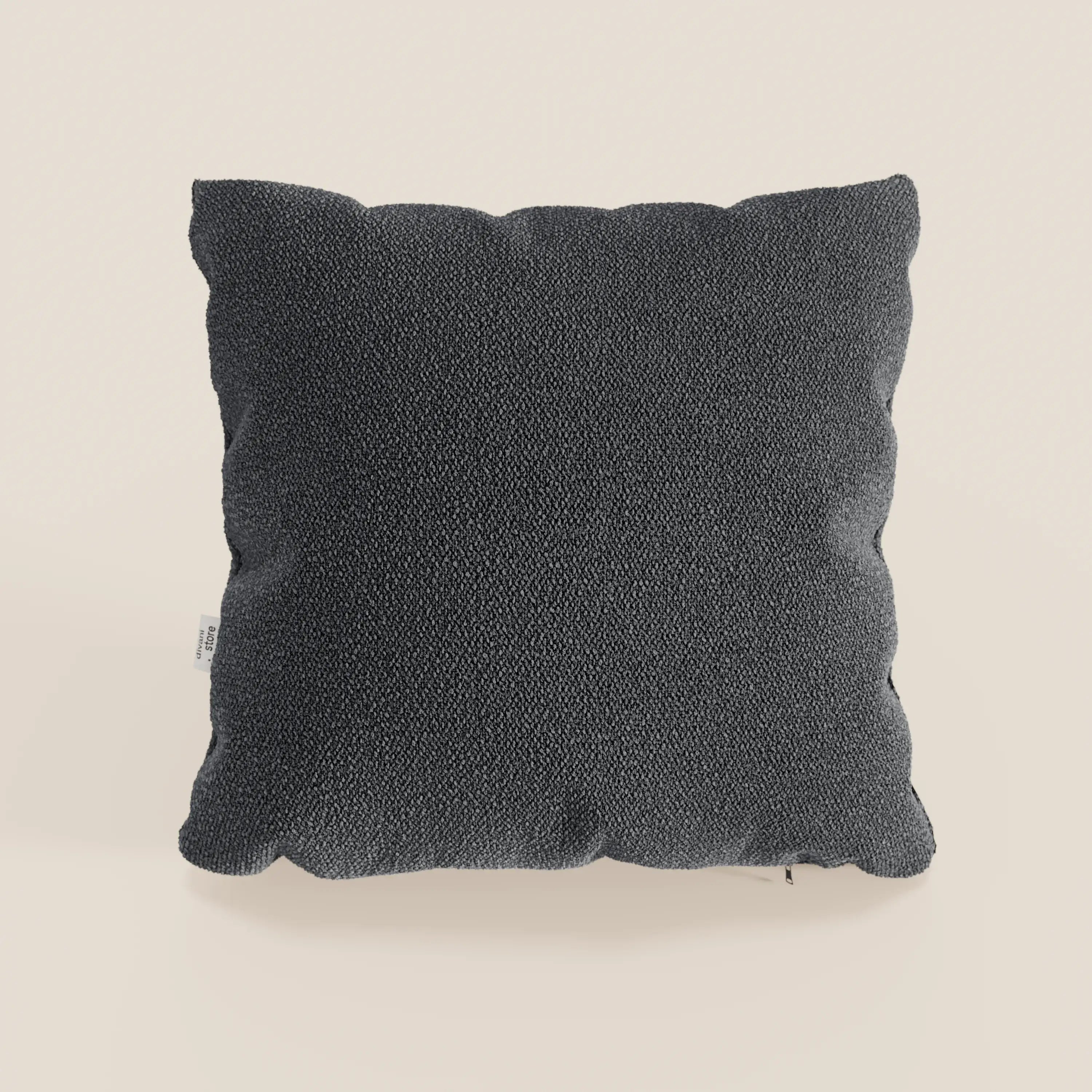 Un cuscino Lambada quadrato grigio antracite di Divani.Store in tessuto smacchiabile e impermeabile tipo cotone naturale si trova in posizione verticale su uno sfondo beige chiaro con pieghe morbide e una piccola etichetta bianca sulla sinistra.