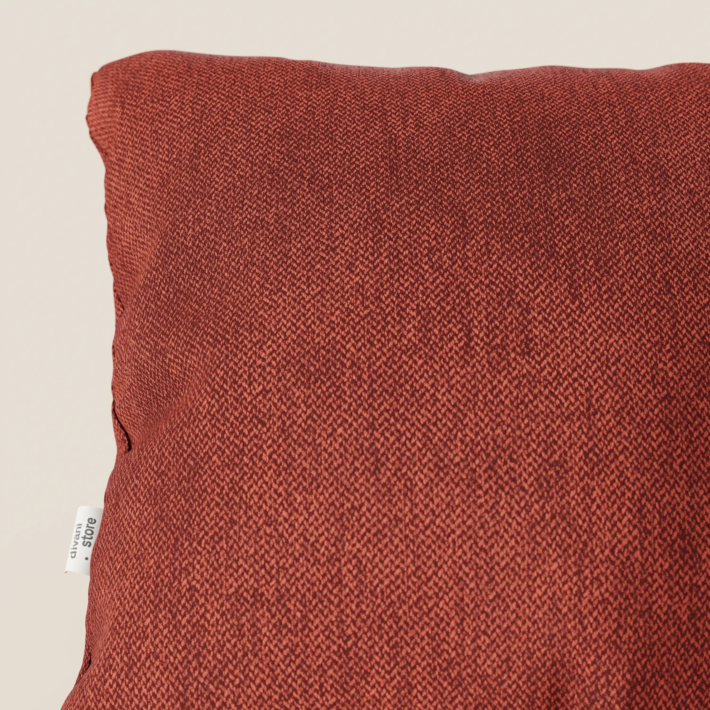 Primo piano della metà superiore del cuscino Sogno di Divani.Store in SMACCHIABILE T05 rosso, caratterizzato da un tessuto strutturato rosso ruggine e da una piccola etichetta bianca sulla cucitura sinistra. Perfetto come cuscino elegante, spicca sul fondo beige chiaro.