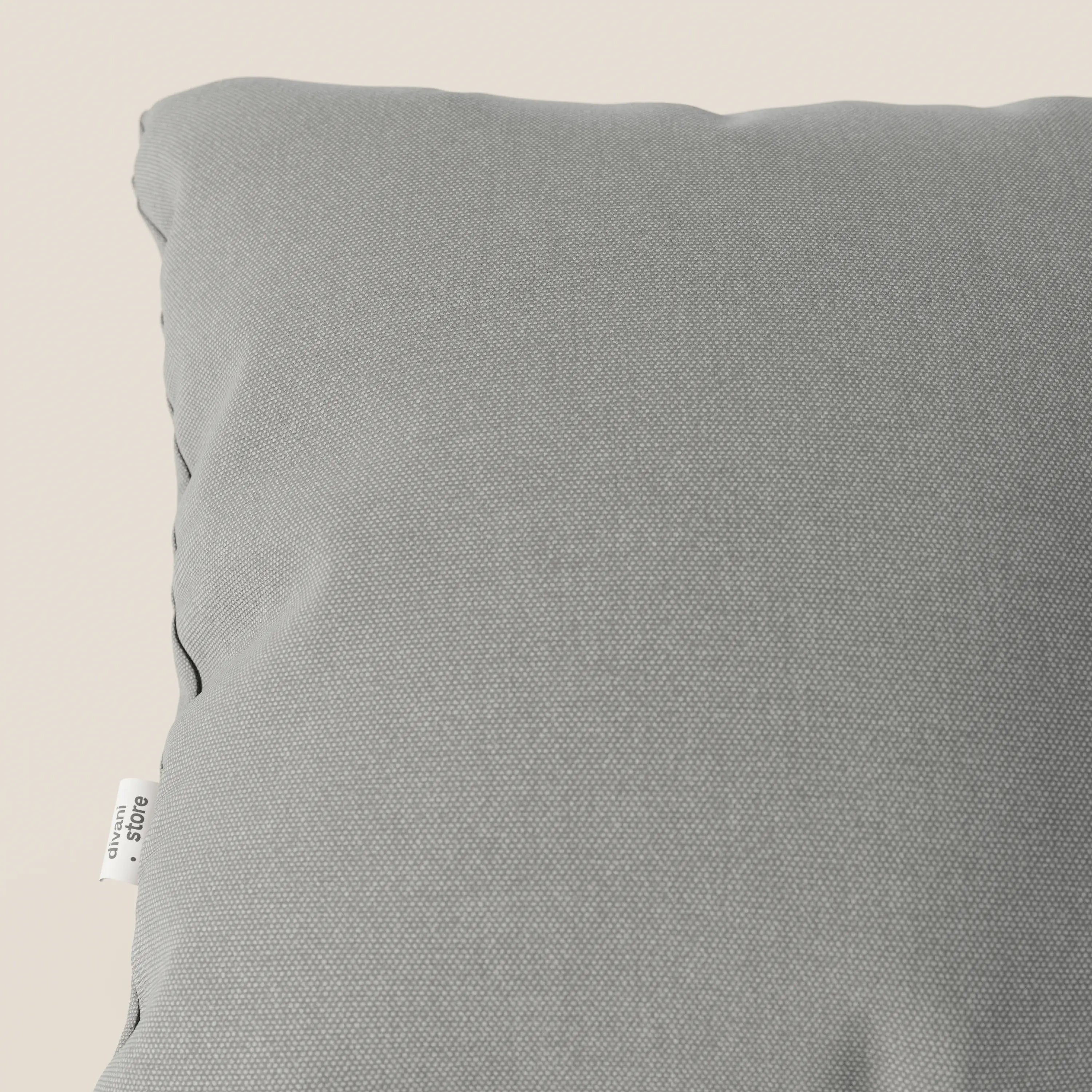 Primo piano del cuscino Honey 45x45 cm di Divani.Store in T13 grigio tessuto simil cotone impermeabile, mostrato in diagonale con l'angolo in alto a sinistra, il lato e il cartellino in tessuto bianco su uno sfondo beige tenue.