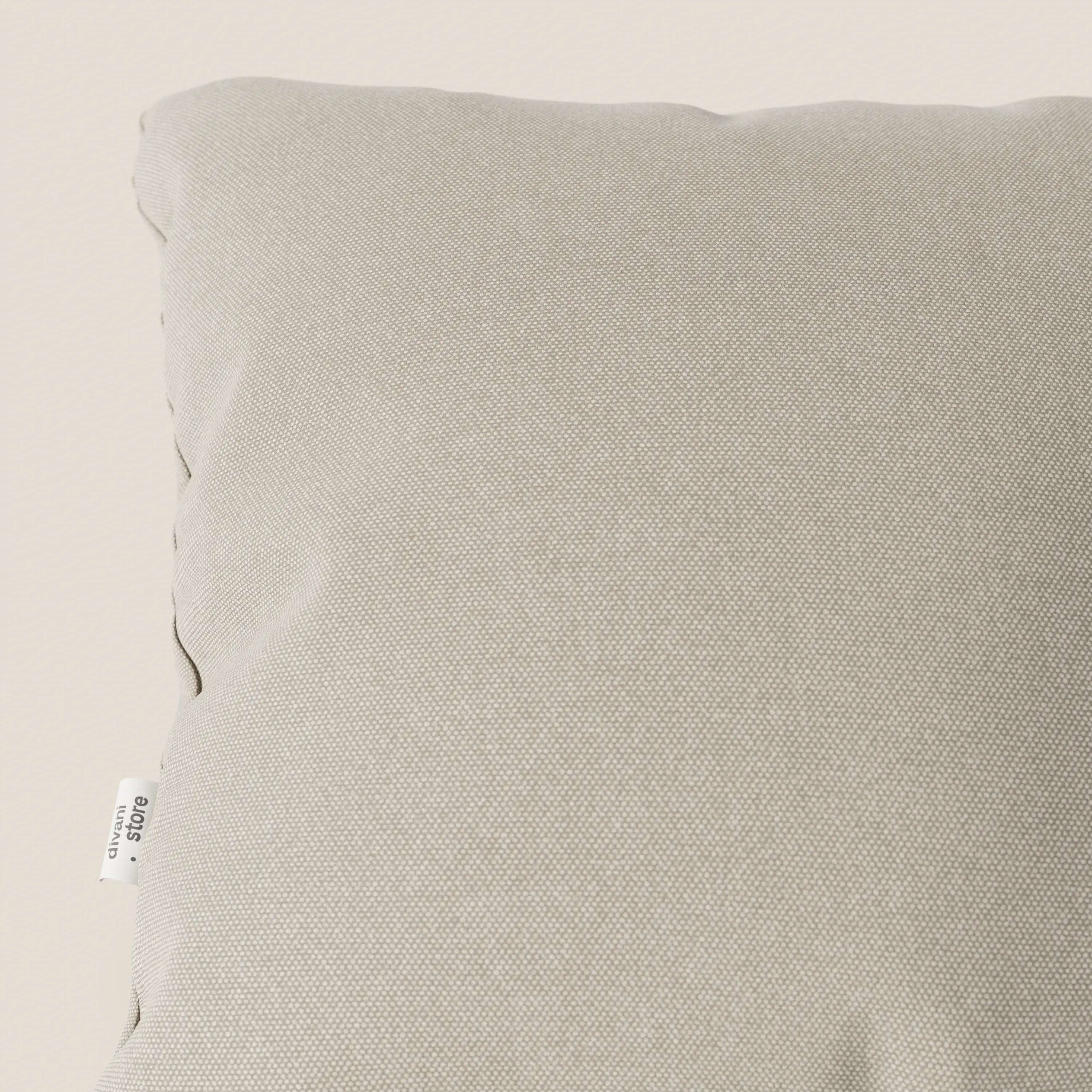 Un primo piano del cuscino Divani.Store Honey 45x45 cm in panna T13 mostra il suo tessuto similcotone impermeabile beige chiaro, leggermente stropicciato e lavabile, con una piccola etichetta laterale bianca su uno sfondo chiaro.