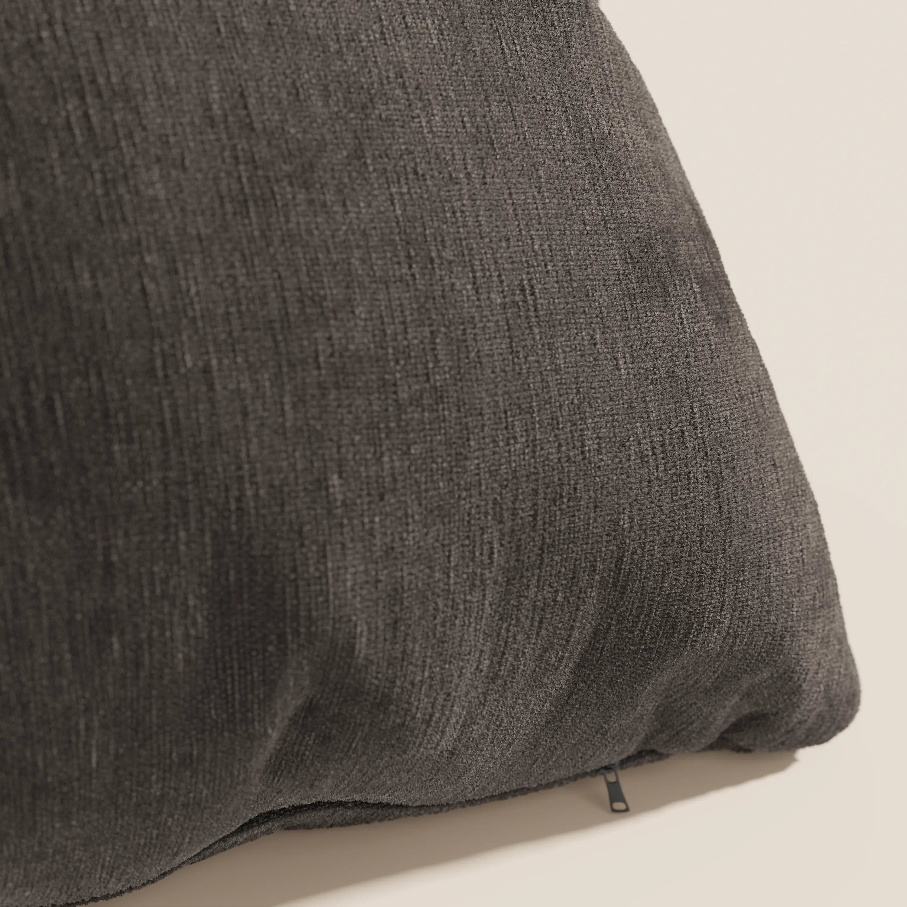 Un primo piano del cuscino Bolero di Divani.Store in grigio scuro mostra il suo tessuto morbido, intrecciato e smacchiabile T31. Il cuscino è posizionato ad angolo su uno sfondo neutro, con una piccola cerniera nera visibile vicino al bordo inferiore.