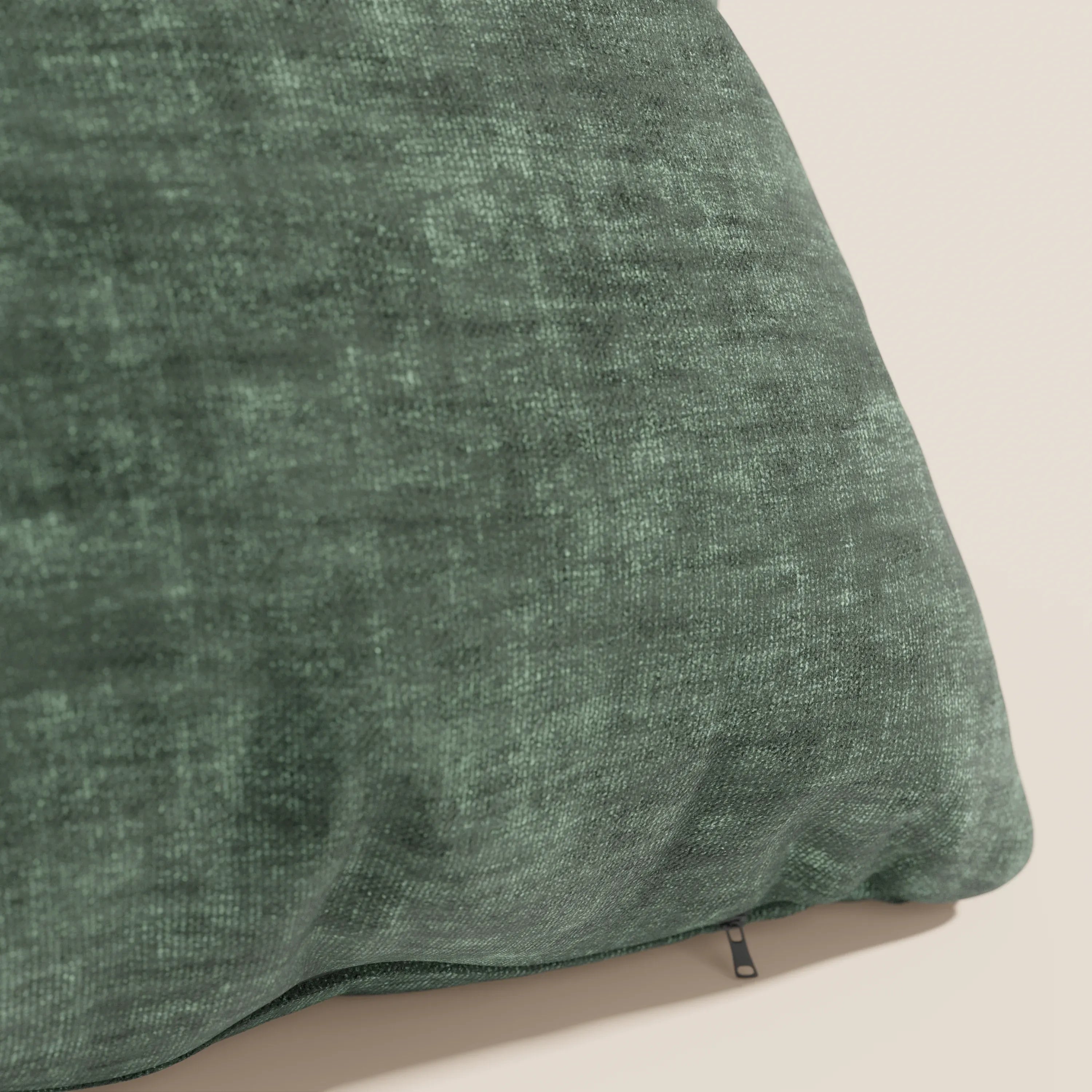 Primo piano del cuscino Sofia di Divani.Store in verde scuro, realizzato in morbido velluto nuvolato (Easy Clean T35) antimacchia e impermeabile, con texture screziata e cerniera inferiore a vista, su una parete in tinta unita beige chiaro.