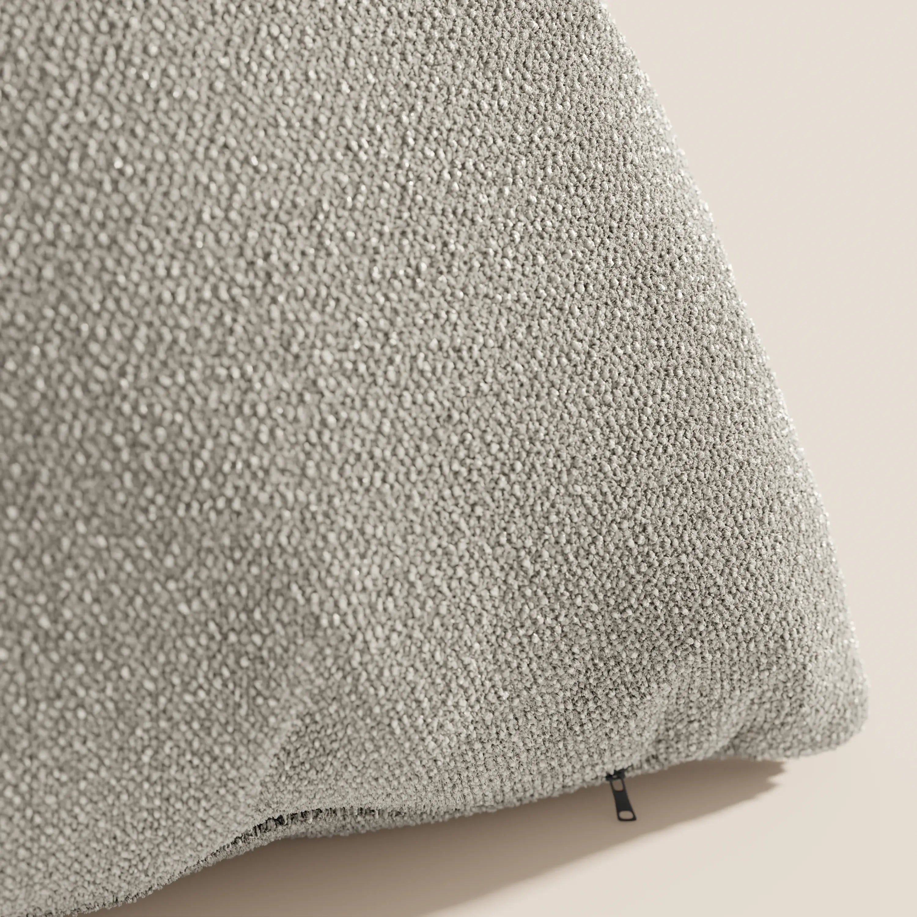 Primo piano del cuscino Lambada di Divani.Store in grigio chiaro, con tessuto simil-cotone (T33) testurizzato, antimacchia e impermeabile, con angolo arrotondato e cerniera nera su un morbido sfondo beige.