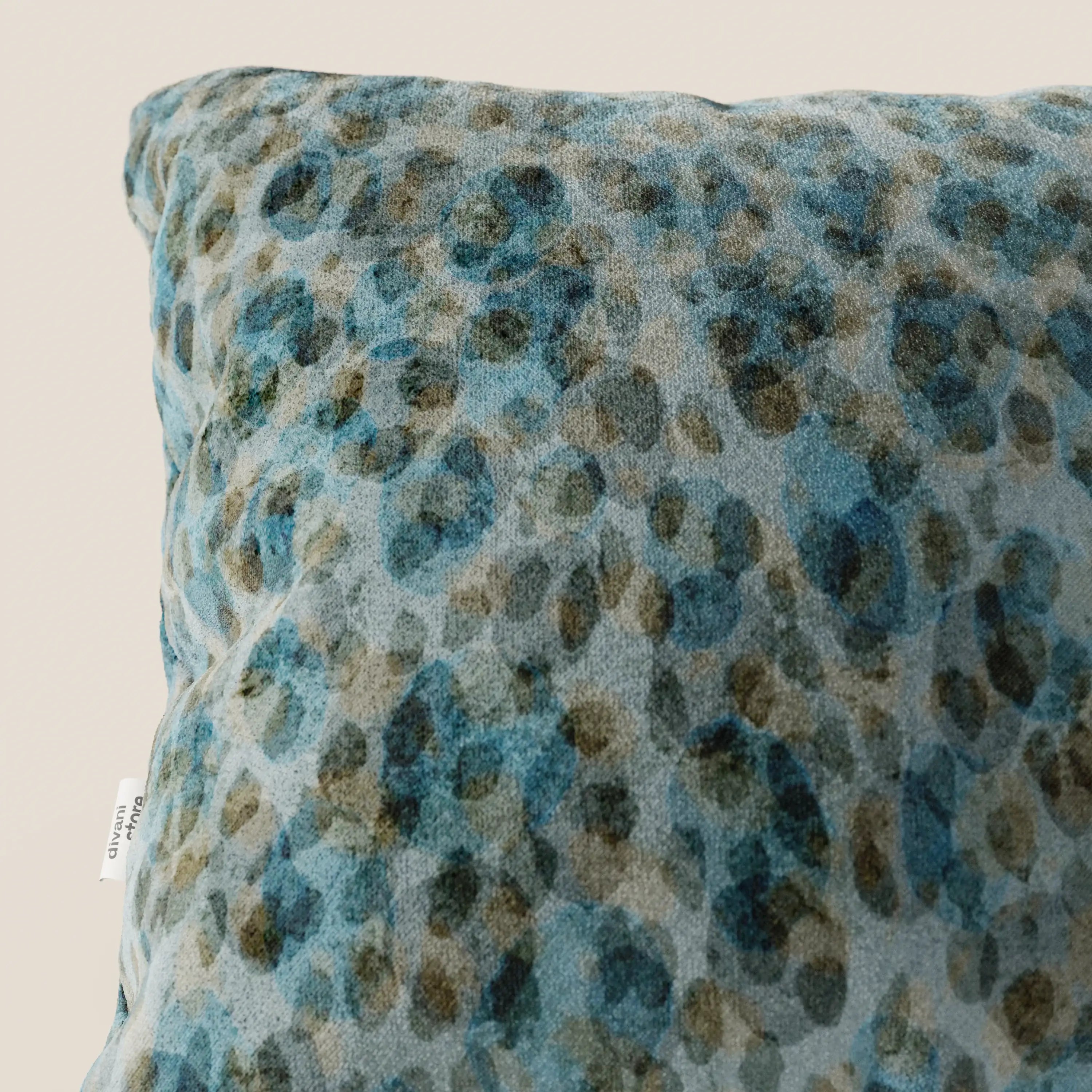 Un primo piano del cuscino Brush 45x45 cm di Divani.Store, realizzato in velluto impermeabile verde acqua, caratterizzato da una texture morbida e da un motivo astratto blu-verde. Sul bordo sinistro compare una piccola etichetta bianca con testo nero.