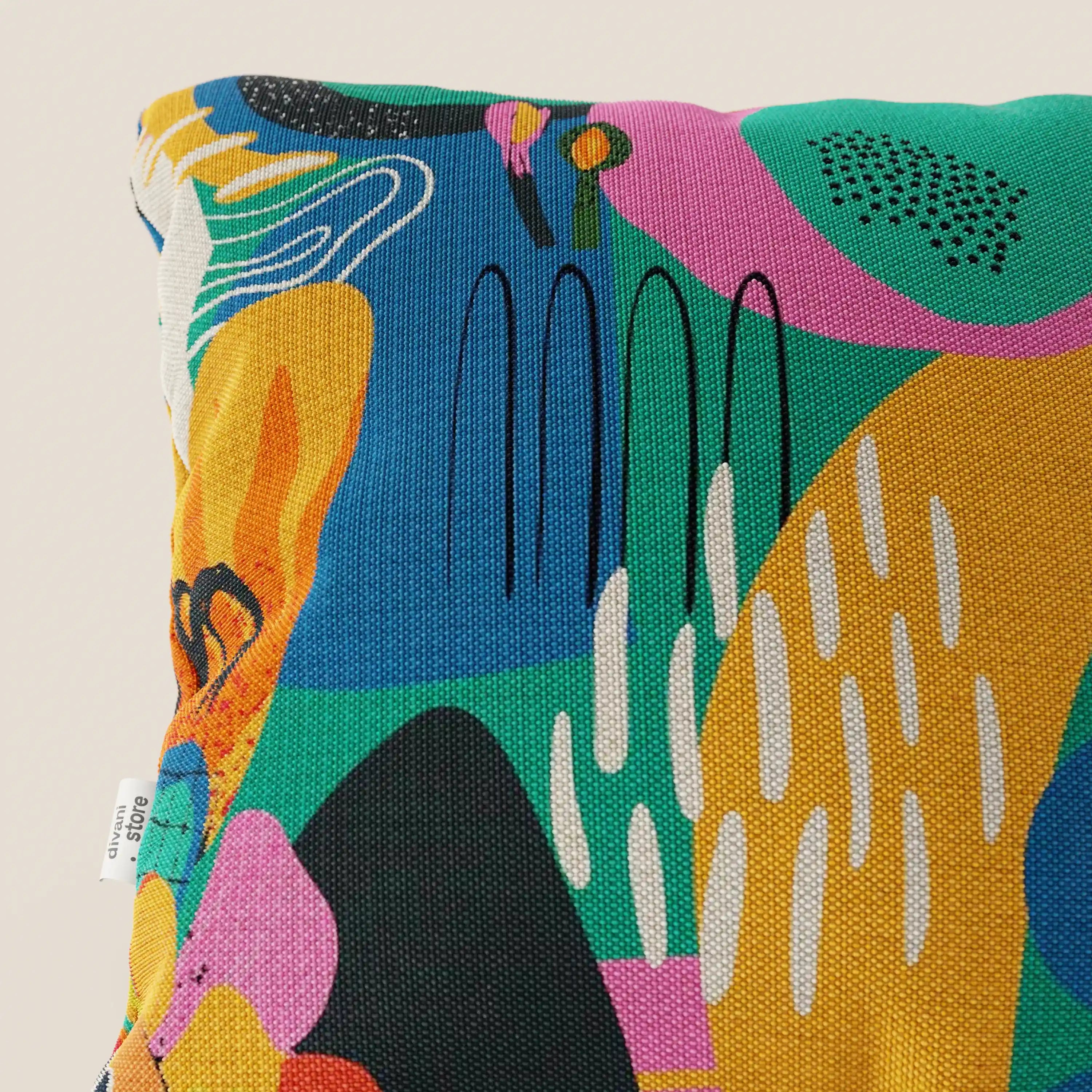 Primo piano del cuscino Joy collezione cuscini di Divani.Store, un cuscino 45x45 cm con stampa astratta multicolore con forme verdi, gialle, arancioni, rosa, nere e bianche su tessuto di cotone impermeabile giallo fresco. Sul bordo sinistro è presente un'etichetta bianca. Sfondo: beige chiaro.