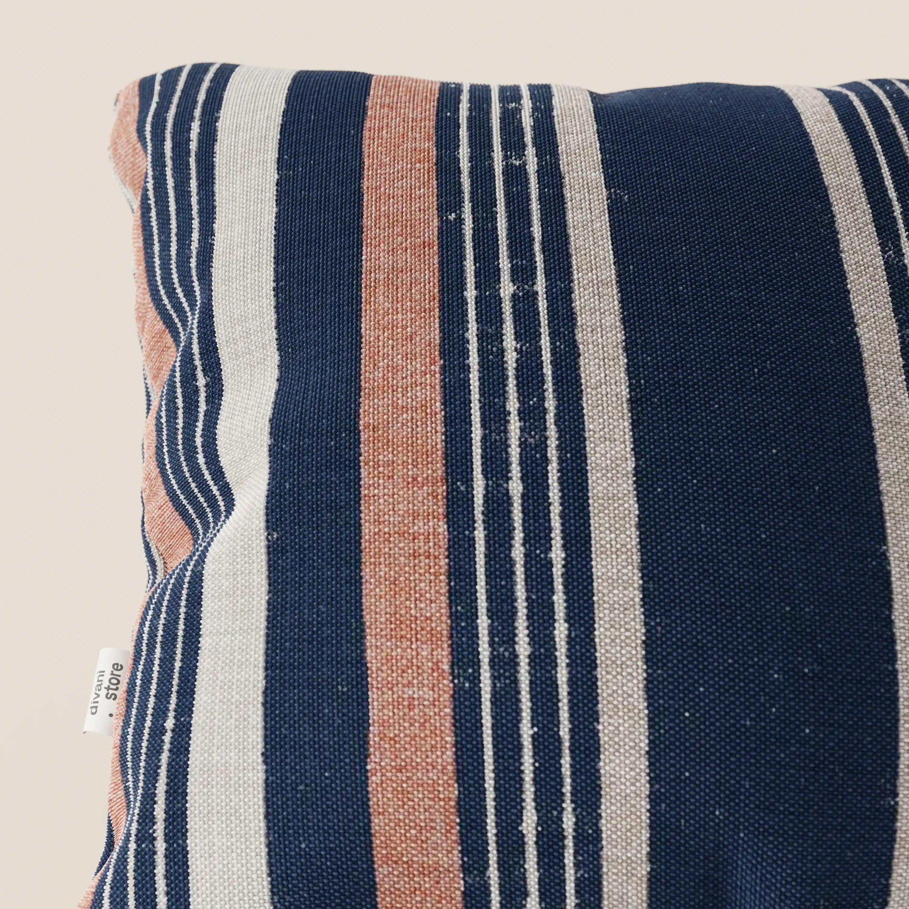 Primo piano dei cuscini a fantasia rigata in tessuto impermeabile di Divani.Store, caratterizzati da ampie strisce blu, bianche e arancioni polverose. Sul bordo sinistro è visibile una piccola etichetta bianca. Lo sfondo è beige chiaro.