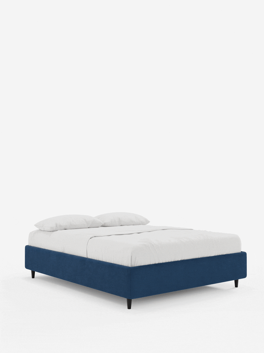 Il letto sommier imbottito ERON con contenitore, rivestito in tessuto impermeabile certificato Oeko-Tex, spicca su uno sfondo bianco, caratterizzato da una base imbottita blu scuro, gambe corte nere, un lenzuolo bianco e due cuscini coordinati.