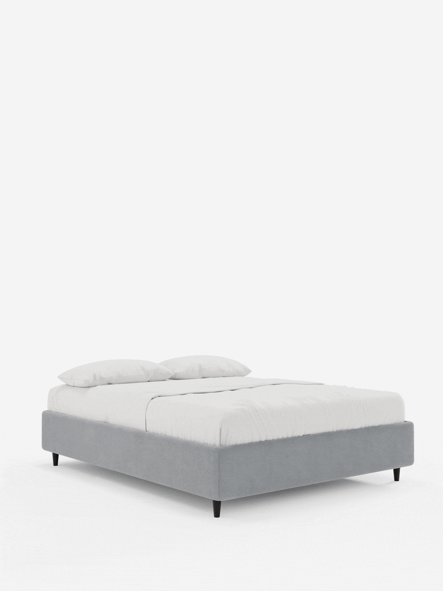 Il letto sommier imbottito ERON con contenitore, realizzato in tessuto impermeabile certificato Oeko-Tex, è un letto a piattaforma rivestito in morbido tessuto grigio, con gambe corte nere affusolate, abbinato a biancheria da letto bianca su uno sfondo grigio chiaro.