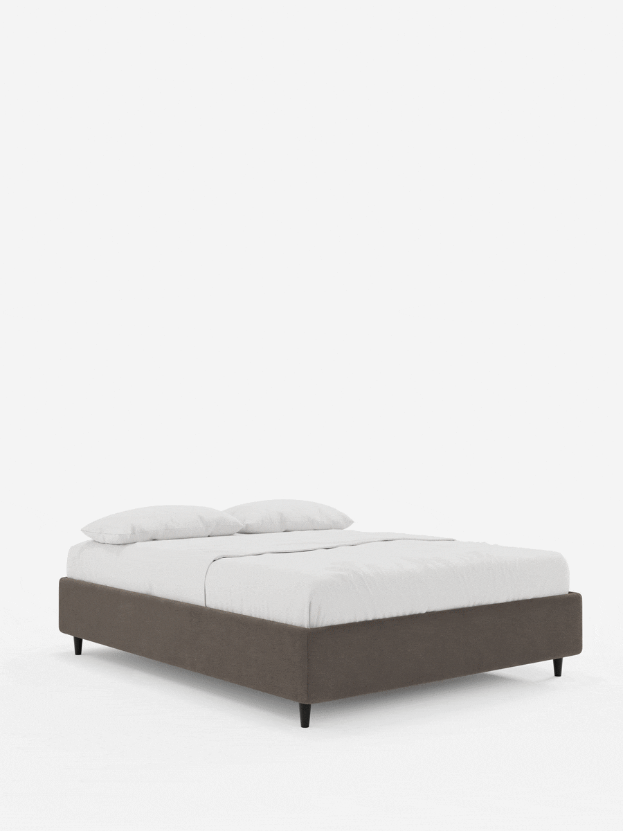 Il letto sommier imbottito ERON con contenitore in tessuto impermeabile certificato Oeko-Tex è un letto imbottito marrone con gambe corte e affusolate di colore scuro, abbinato a lenzuola bianche immacolate e due cuscini coordinati su uno sfondo bianco a tinta unita.