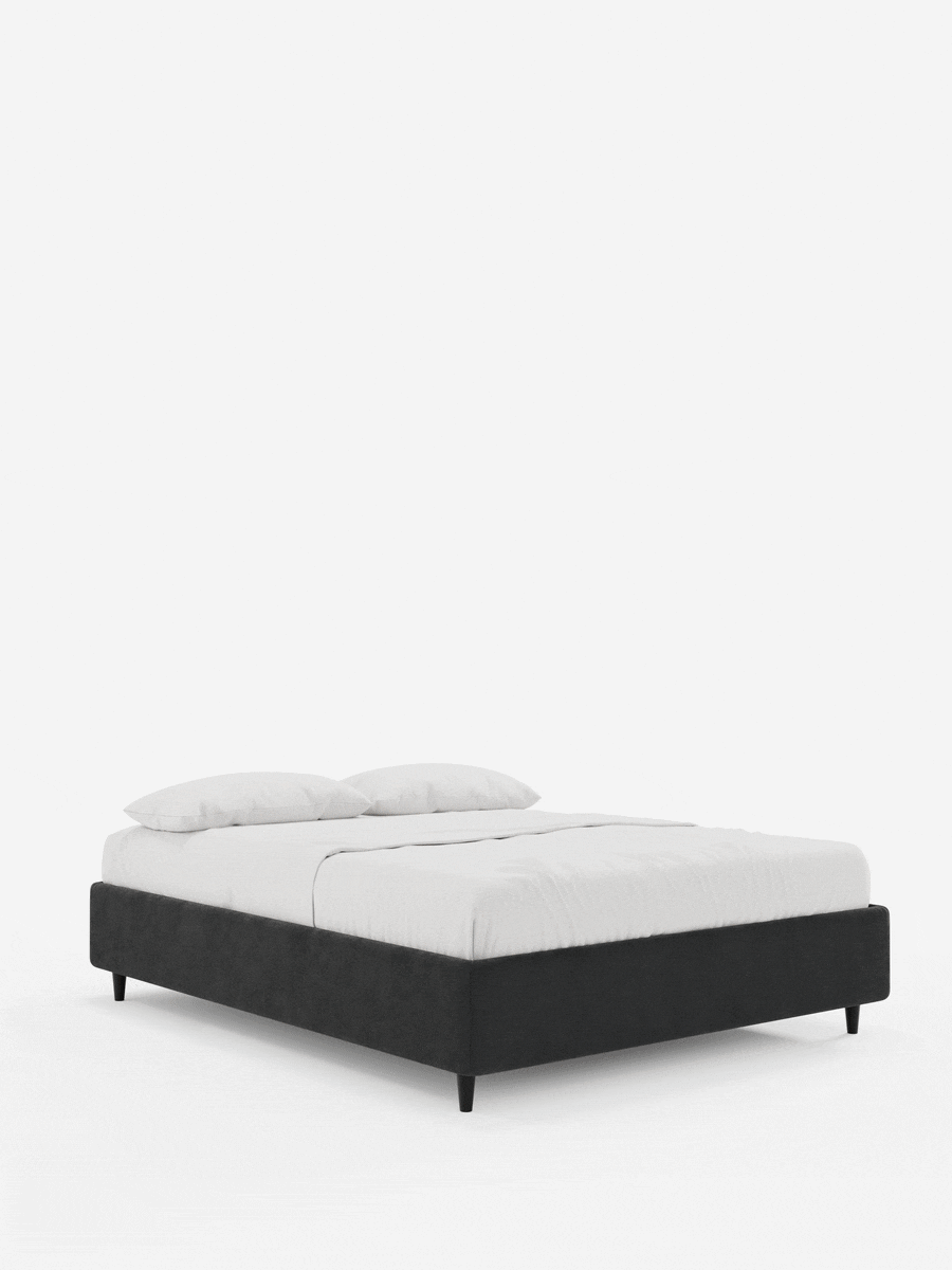 Il letto sommier imbottito ERON con contenitore, rivestito in tessuto impermeabile certificato Oeko-Tex, è un letto dal profilo basso in tessuto grigio scuro, con gambe corte nere, rivestito con lenzuola bianche immacolate e due cuscini su uno sfondo bianco sporco.