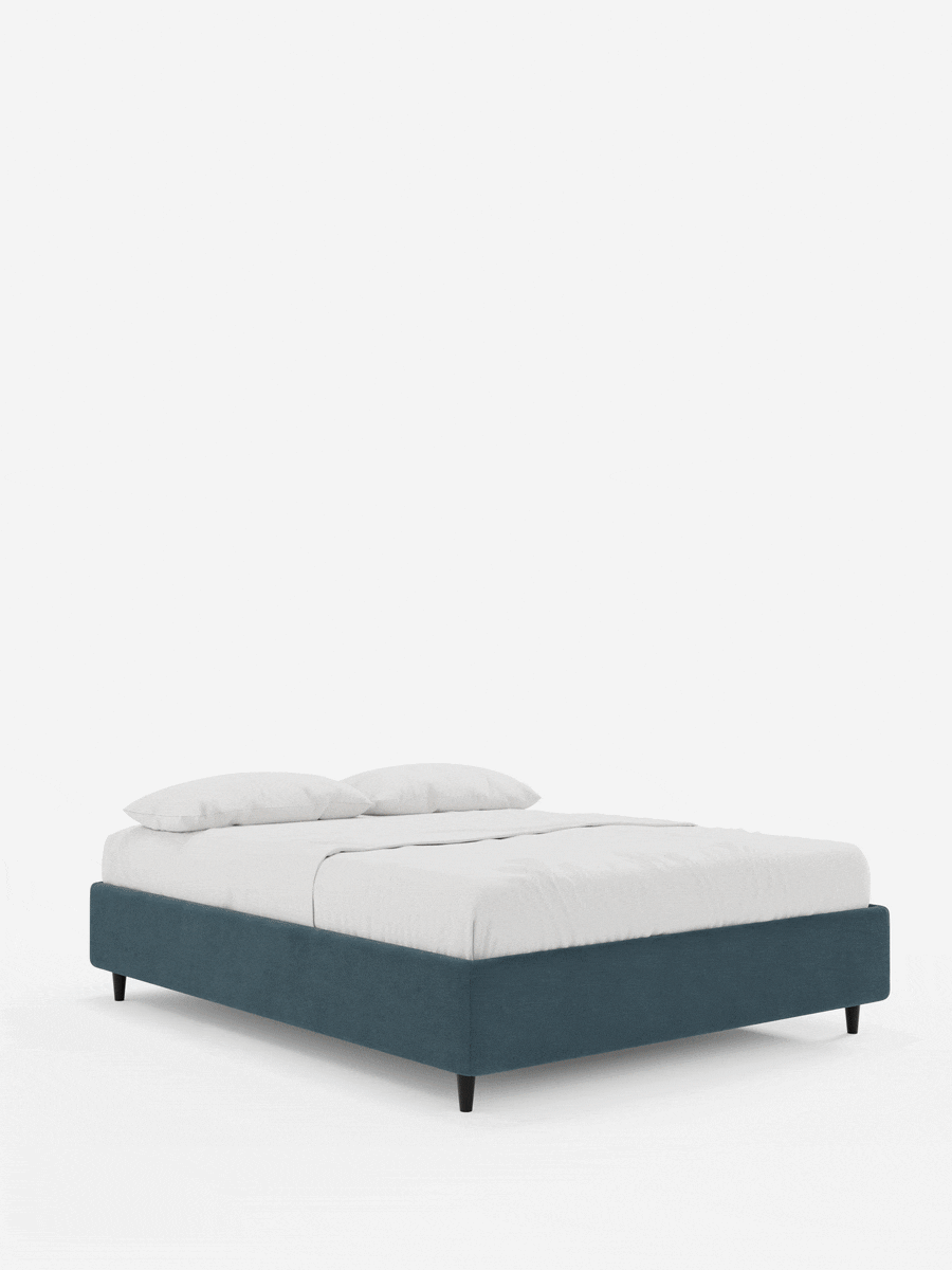 Il letto sommier imbottito ERON con contenitore in tessuto impermeabile certificato Oeko-Tex è un letto dal profilo basso, rivestito in tessuto blu scuro, con biancheria da letto bianca, due cuscini e gambe corte e affusolate di colore scuro, che risaltano su uno sfondo bianco.