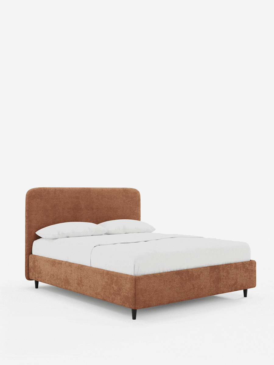 Un letto KAELOS in stile minimal, imbottito in ciniglia color ruggine su sfondo bianco, caratterizzato da una testiera arrotondata, sottili gambe nere affusolate, lenzuola bianche e due cuscini.