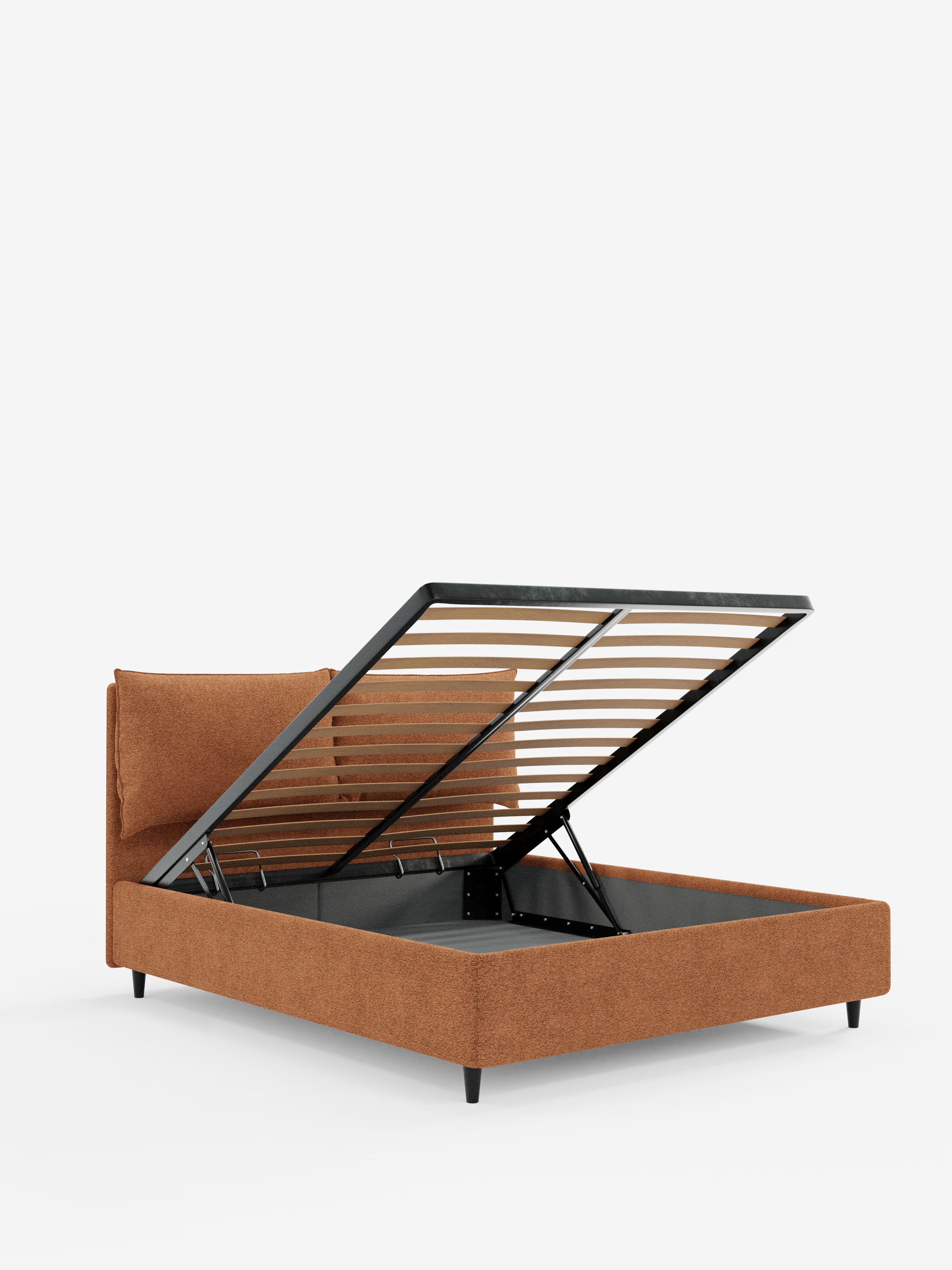 Un letto imbottito contemporaneo ZAYRON con contenitore di Letti.store, realizzato in tessuto color ruggine certificato Oeko-Tex®. Caratterizzato da una testiera con morbidi cuscini regolabili e da gambe affusolate nere, il letto è raffigurato con la rete a doghe sollevata per rivelare l'ampio spazio di contenimento nascosto al di sotto, su uno sfondo bianco a tinta unita.