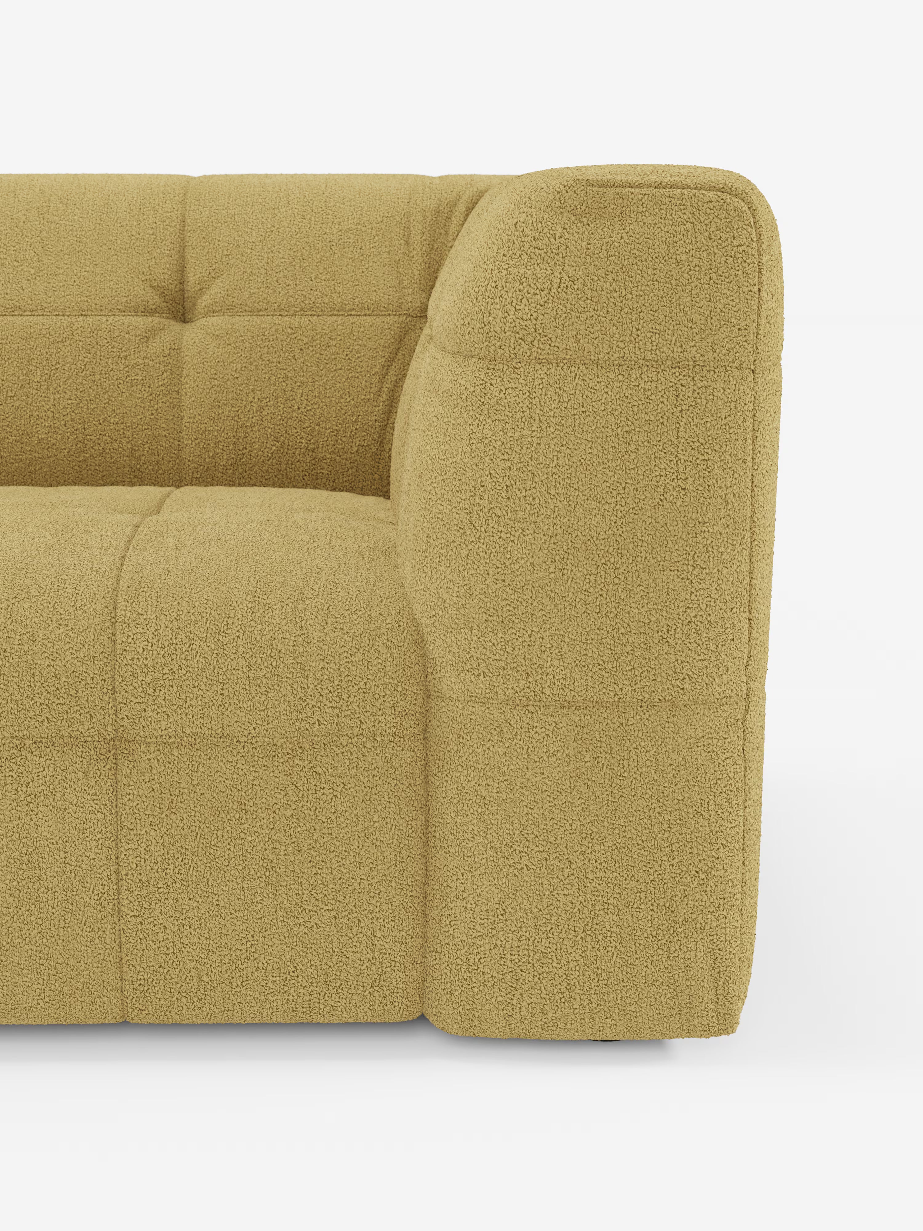 Vista parziale di un divano moderno rivestito in tessuto testurizzato color beige chiaro, che mostra il bracciolo e la seduta. Questo divano moderno presenta una profonda trapuntatura a quadrati e bordi morbidi e arrotondati, che risaltano su uno sfondo bianco a tinta unita.