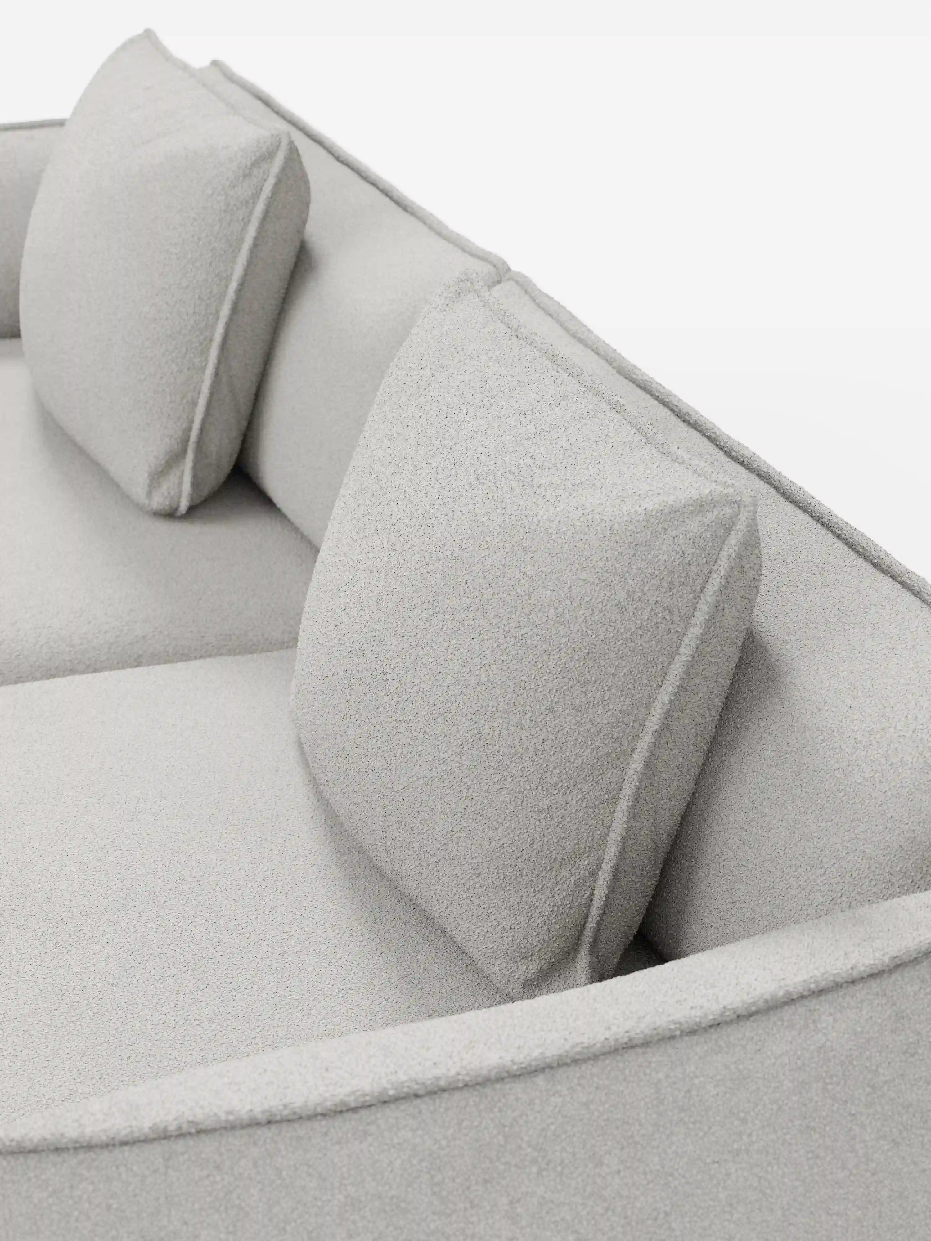 Un primo piano del divano Nimbus a 3 posti, modulare e componibile da 250 cm di Divani.Store, rivestito in tessuto smacchiabile grigio chiaro effetto cotone T33, mette in risalto due cuscini quadrati abbinati. Il tessuto morbido e dalla trama particolare, insieme all’illuminazione brillante, esaltano il design pulito, minimalista e moderno del divano.