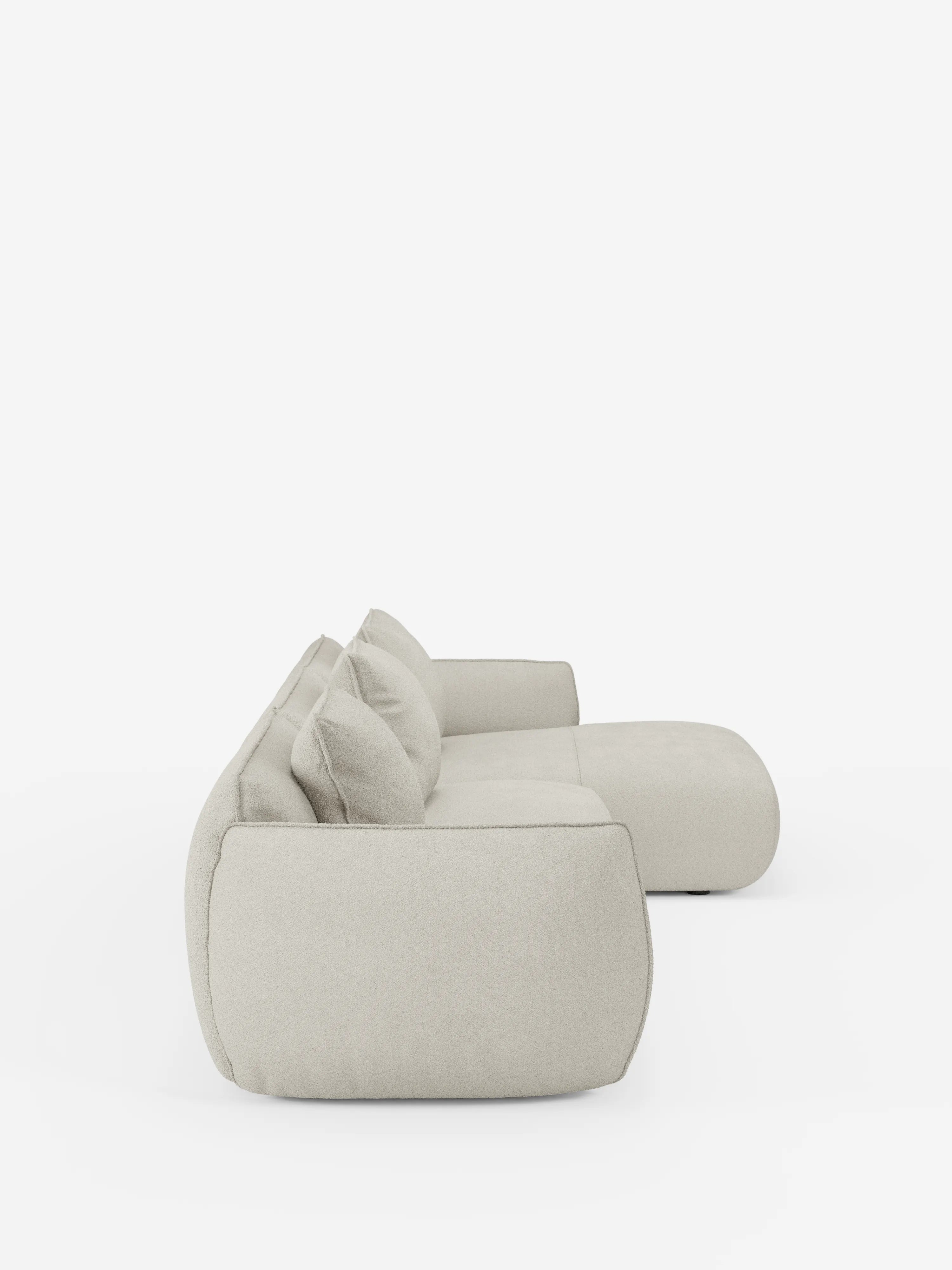 Vista laterale del divano angolare con penisola componibile Nimbus di Divani.Store, caratterizzato da un design moderno e minimalista in beige chiaro. Il divano è rivestito in morbido tessuto effetto cotone antimacchia (T33) con bordi lisci e arrotondati. Due cuscini coordinati poggiano sulla seduta. Lo sfondo bianco a tinta unita esalta la trama morbida e raffinata di questo complemento d’arredo contemporaneo.