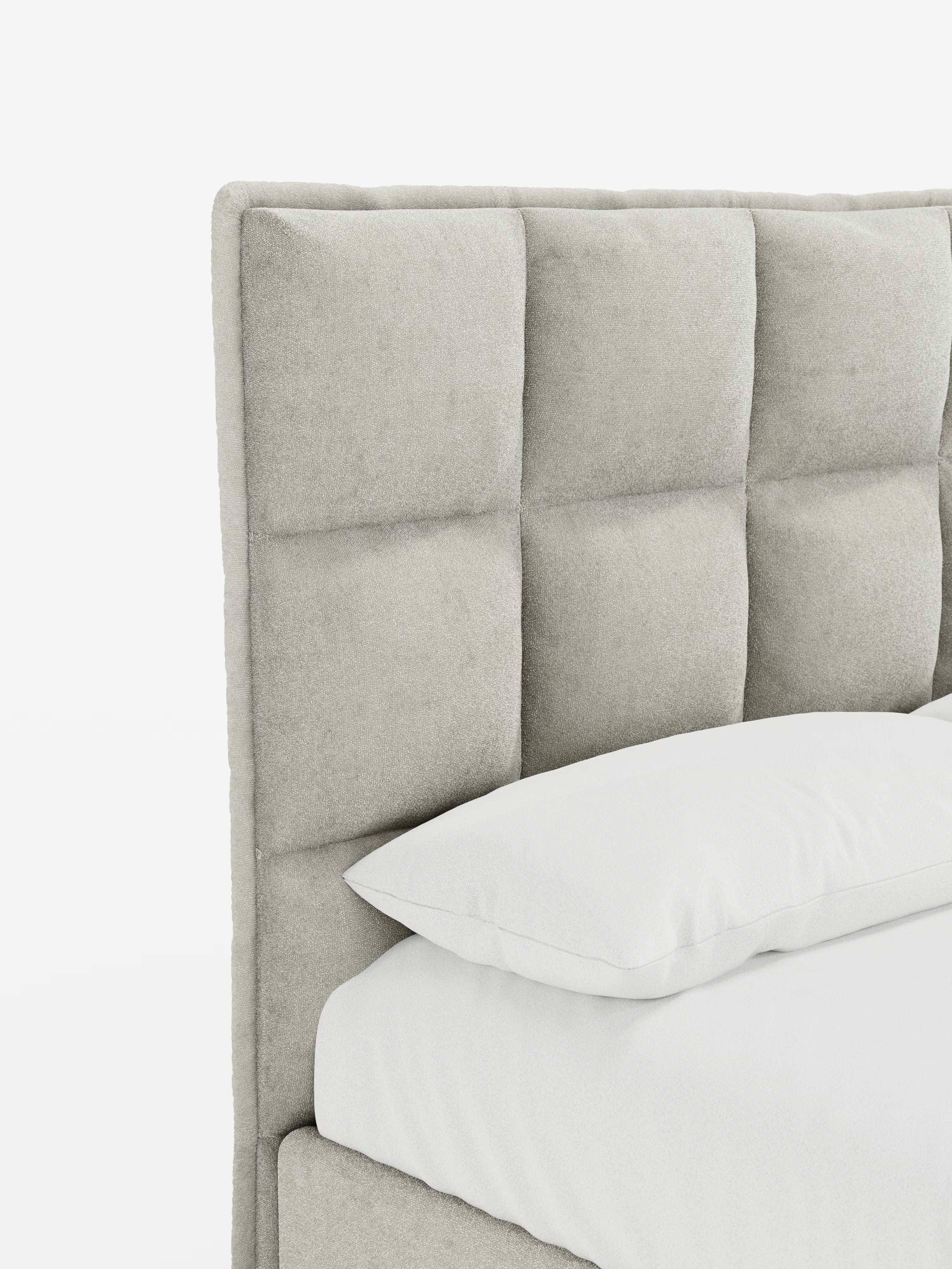 Primo piano del letto AURO di Letti.store, caratterizzato da una testiera alta imbottita di colore grigio chiaro con trapuntatura a quadrati in ciniglia Oeko-Tex®, che richiama il classico effetto capitonné. Un cuscino bianco e una parte del materasso bianco mettono in risalto la morbida trama e il design contemporaneo e minimalista. Lo sfondo è bianco a tinta unita.