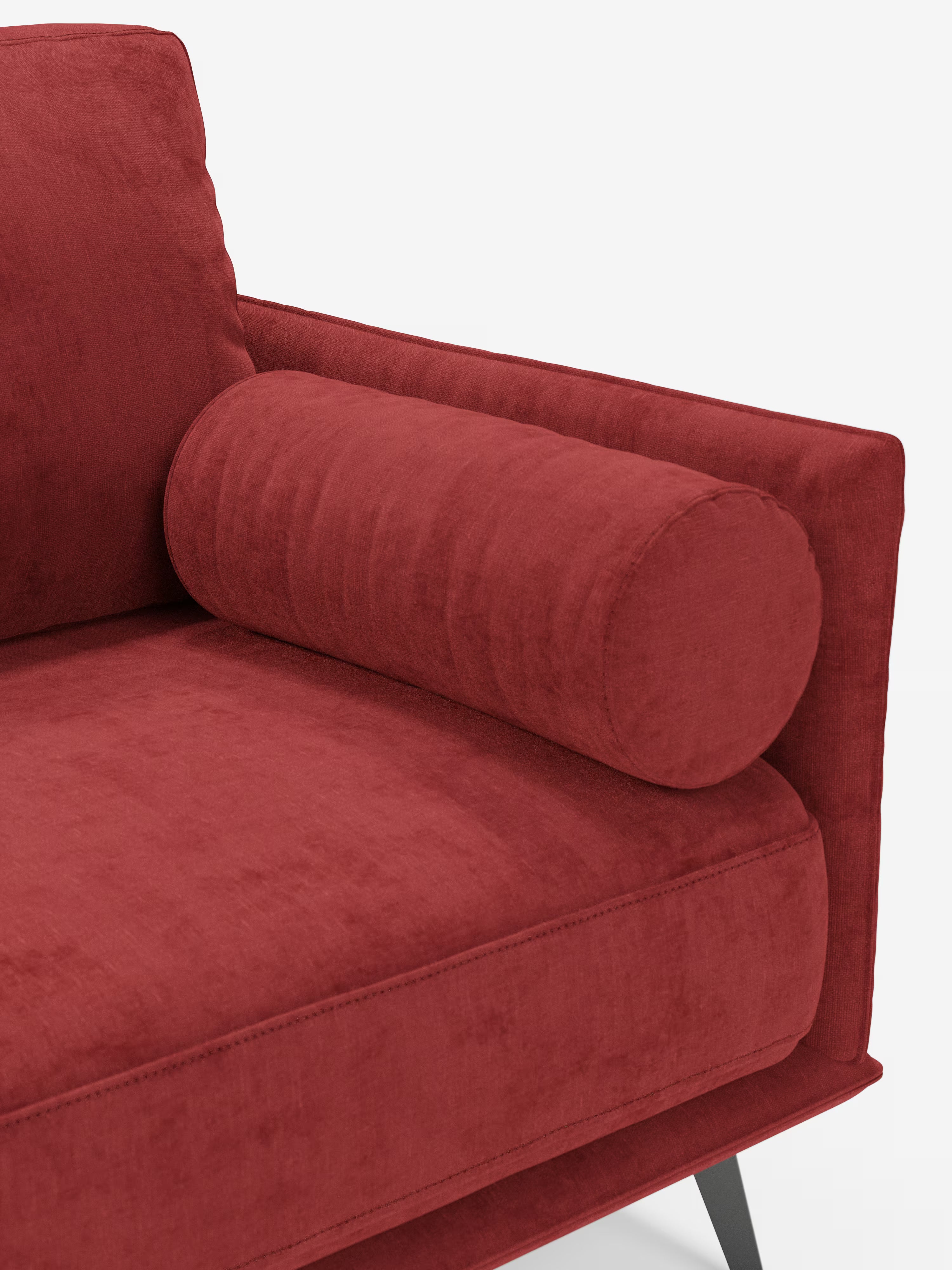 Un primo piano del moderno divano lineare a 2 posti Basalto di Divani.Store, in vivace velluto rosso, mette in risalto un cuscino cilindrico coordinato appoggiato sulla seduta vicino al bracciolo. Questo divano dal design minimalista presenta morbidi cuscini, una silhouette squadrata ed eleganti gambe angolari in metallo nero — il tutto realizzato in tessuto smacchiabile T31 — che risaltano su uno sfondo bianco candido.