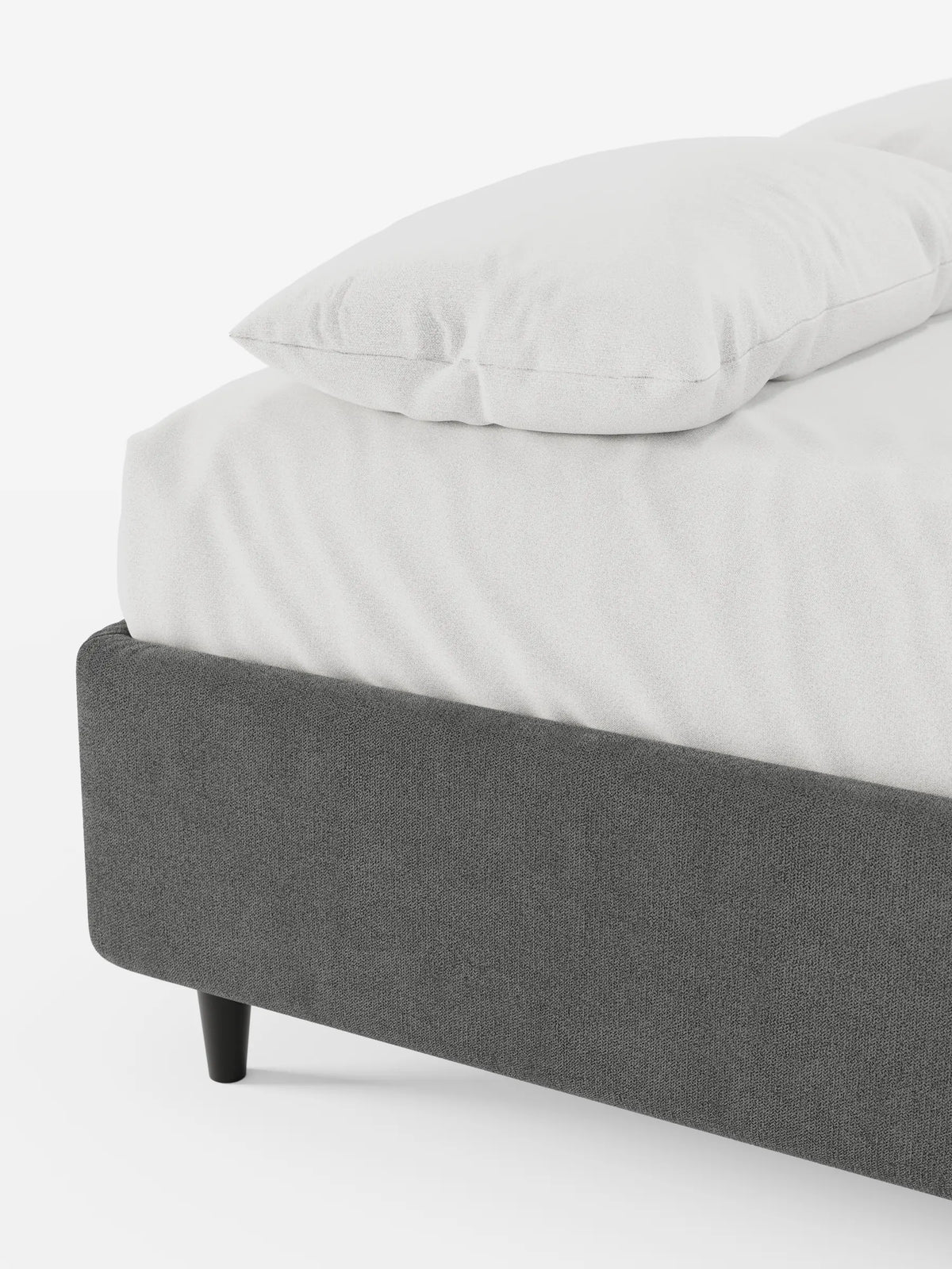 Un primo piano dell'angolo di un letto sommier imbottito ERON grigio scuro con vano contenitore in tessuto impermeabile certificato Oeko-Tex, su gambe corte nere, rivestito con un lenzuolo bianco e un cuscino su uno sfondo bianco sporco.