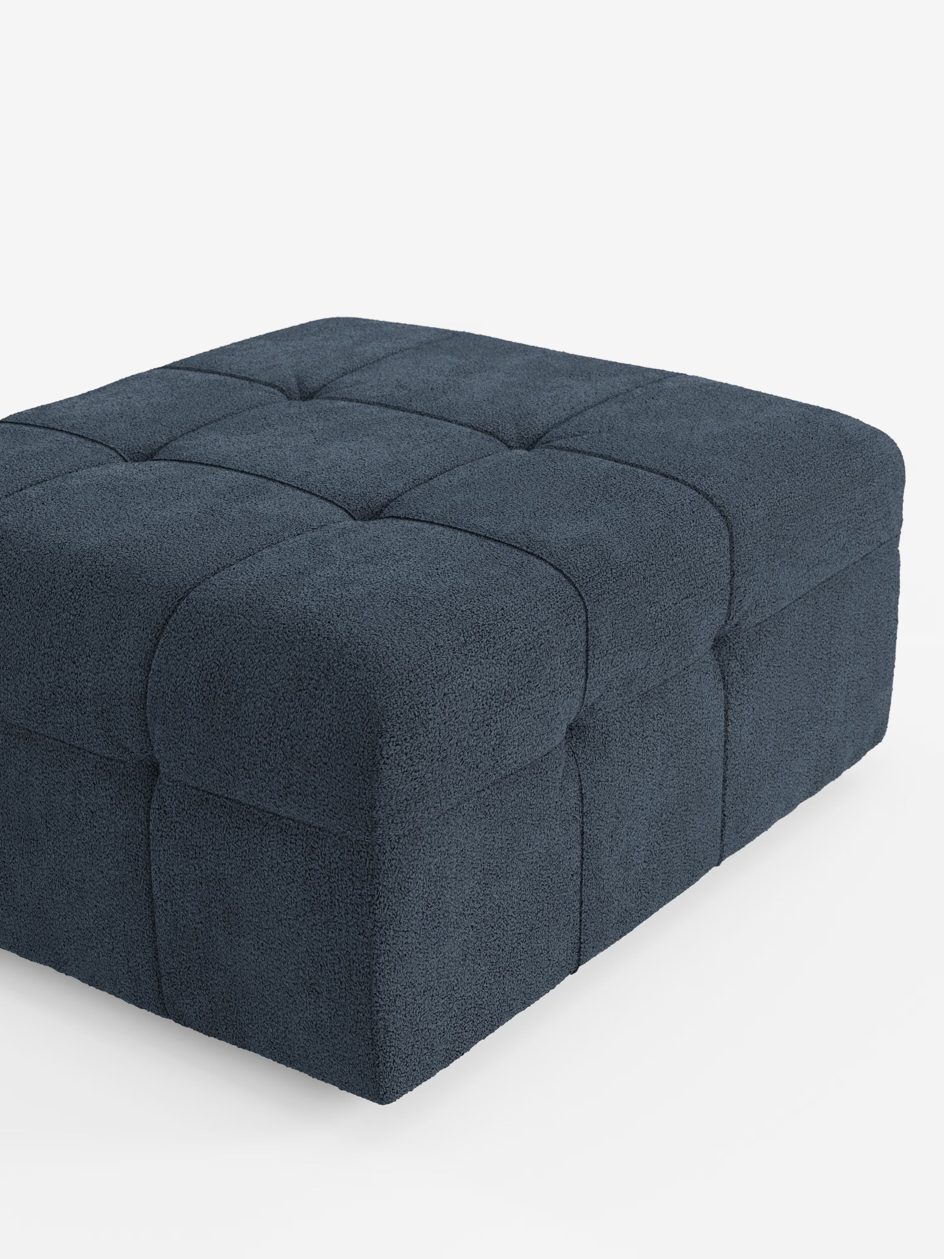 Un pouf e modulo Mosaic di Divani.Store, spazioso e di forma quadrata, realizzato in bouclé, che misura 96x77 cm ed è rivestito in tessuto T21 blu intenso e resistente alle macchie, con un motivo capitonné trapuntato suddiviso in nove quadrati sulla parte superiore e sui lati, è esposto su uno sfondo bianco.