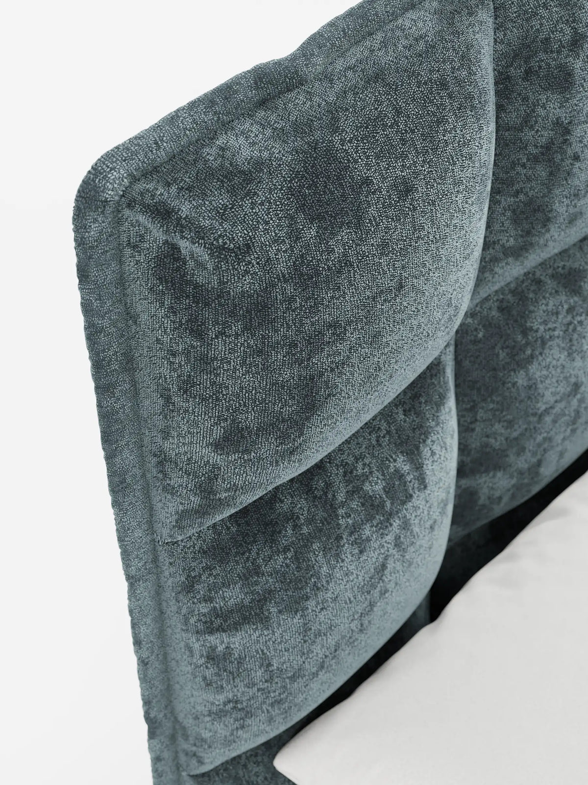 Primo piano del letto imbottito AURO con contenitore e testiera alta capitonné in ciniglia Oeko-Tex®, caratterizzato da una morbida testiera quadrata capitonné in grigio-verde acqua, accanto a un cuscino bianco su uno sfondo chiaro e uniforme.