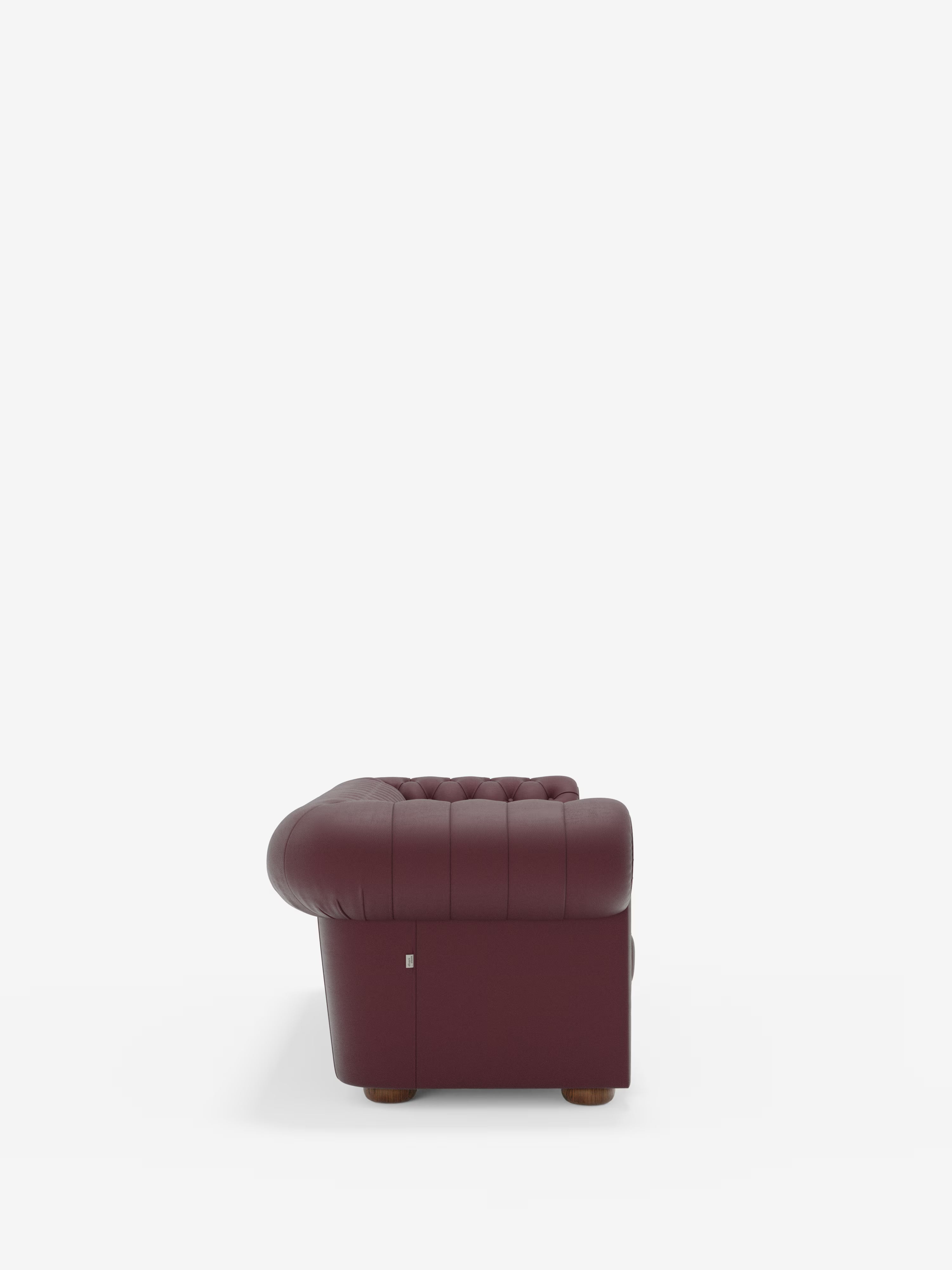 Vista laterale del Chester – Divano Chesterfield in vera pelle spessorata a 2 e 3 posti di Divani.Store, qui raffigurato in una tonalità bordeaux scuro. Questa lussuosa poltrona capitonné presenta braccioli e schienale arrotondati, nel classico stile Chesterfield, e poggia su piccoli piedini in legno su uno sfondo bianco a tinta unita.