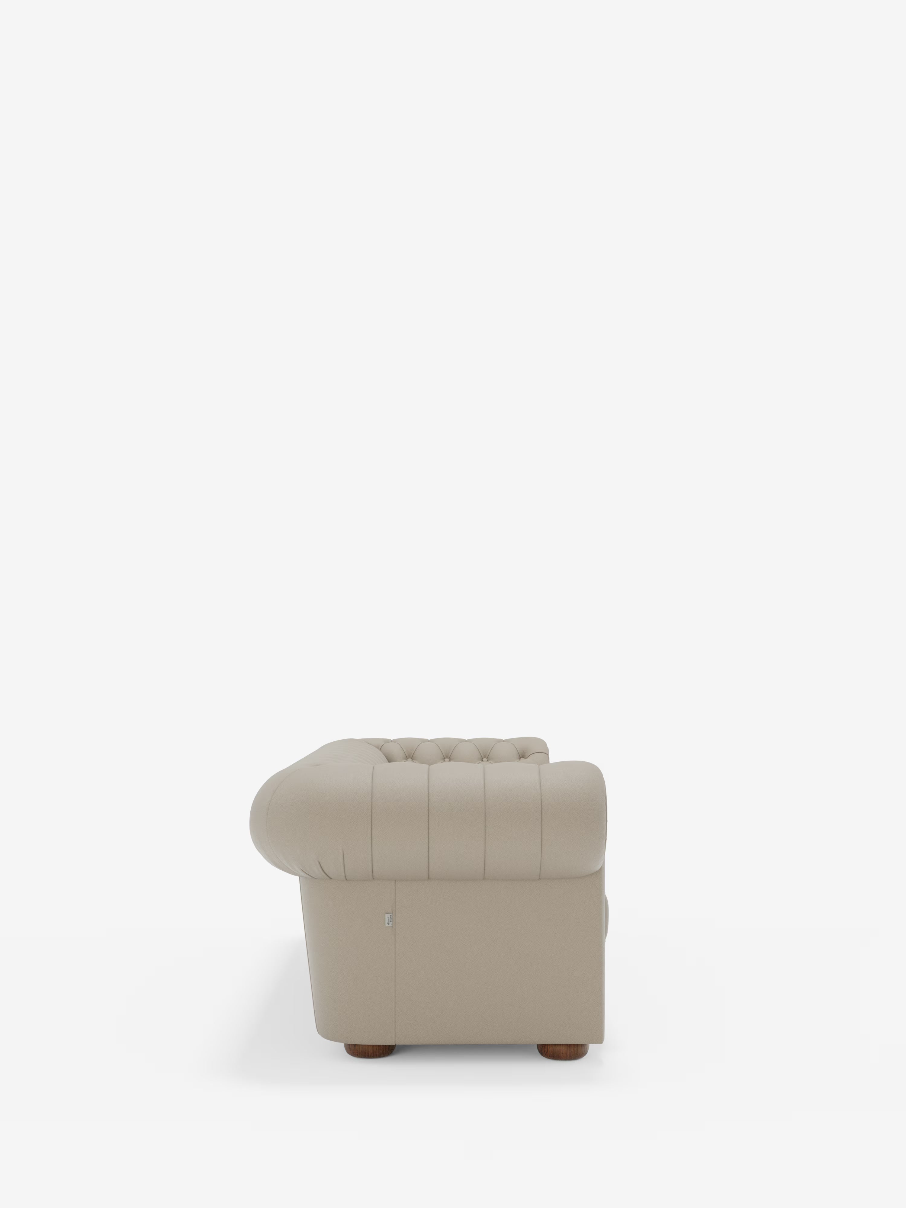 Vista laterale della “Chester 4 Posti 3 sedute in Vera Pelle – Chesterfield Capitonné Artigianale” di Divani.Store, una poltrona beige capitonné con braccioli arrotondati e piedini in legno, su uno sfondo bianco a tinta unita. La poltrona presenta cuciture a vista sui braccioli e sullo schienale, vanta una finitura liscia in vera pelle e incarna la classica maestria artigianale made in Italy.