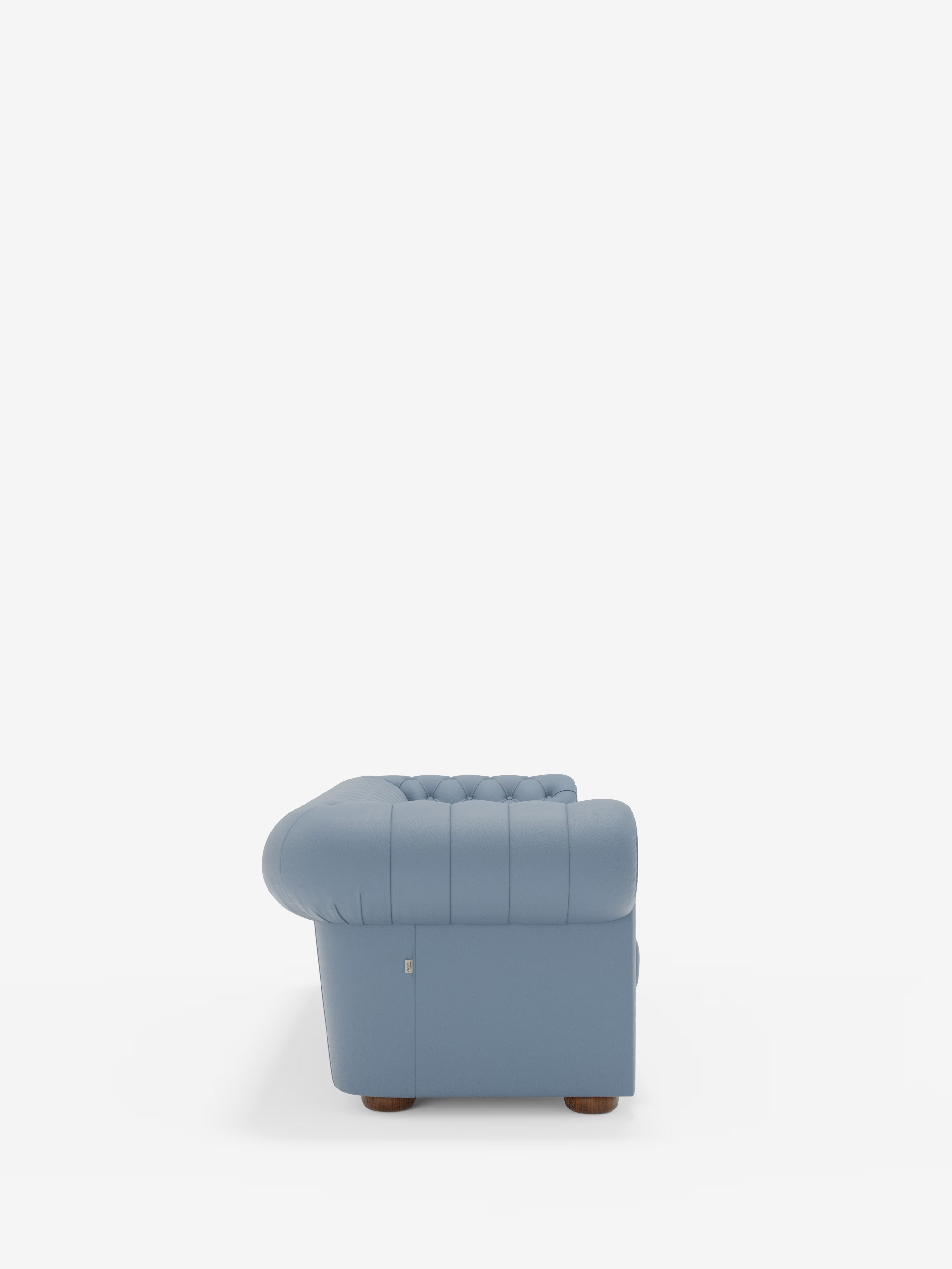 Vista laterale del divano Chester 4 Posti 3 sedute in vera pelle – Chesterfield capitonné artigianale di Divani.Store in azzurro, su uno sfondo bianco a tinta unita. Rivestito in vera pelle e realizzato in Italia, questo divano presenta braccioli capitonné, cuciture a vista e gambe corte e arrotondate in legno, posizionate con discrezione alla base.