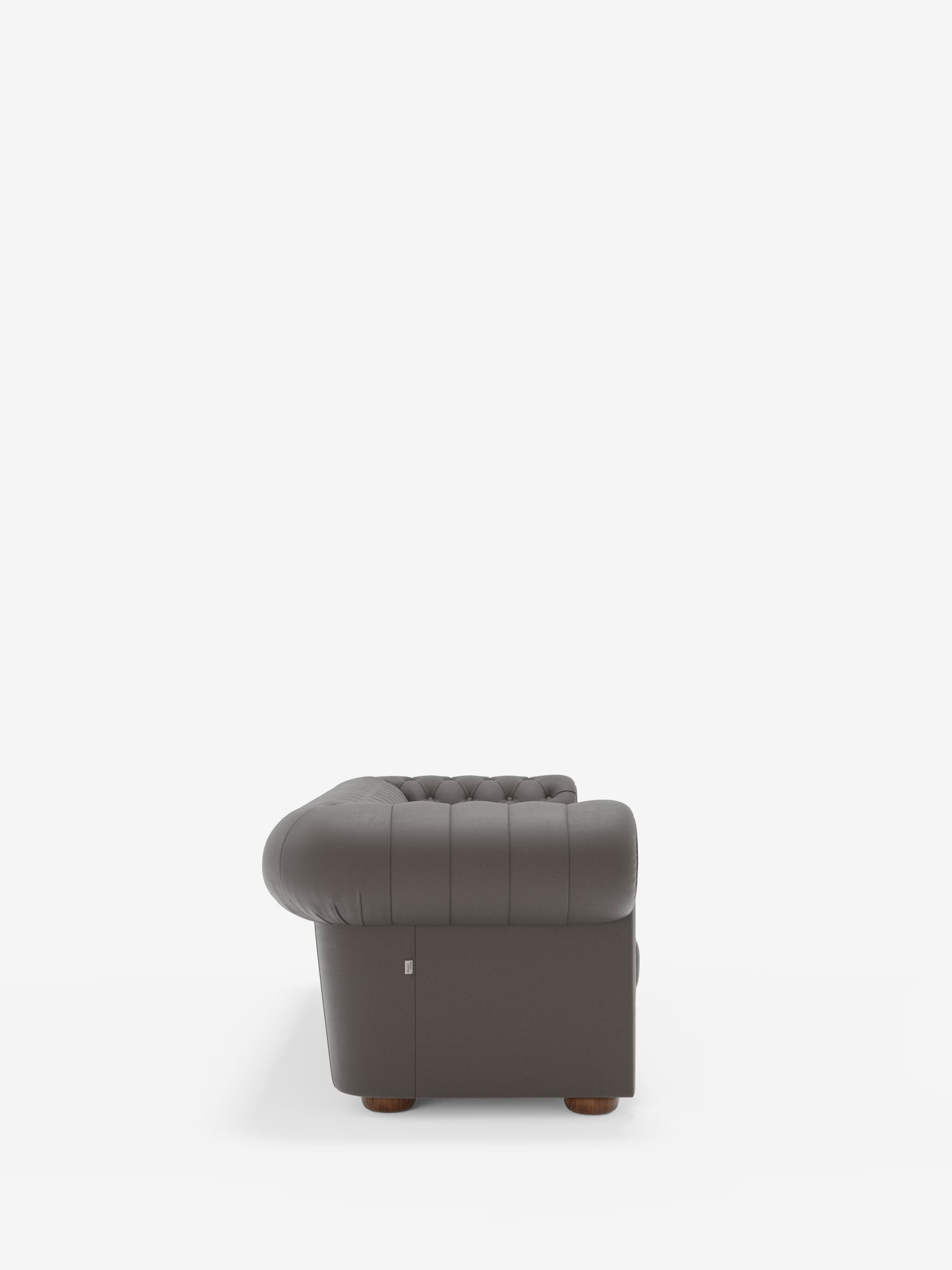 Un divano Chester 4 Posti 3 sedute in vera pelle, colore grigio scuro – Chesterfield Capitonné Artigianale di Divani.Store, visto di profilo, mette in risalto i braccioli e lo schienale arrotondati, le gambe in legno e una piccola etichetta metallica nell’angolo inferiore, su uno sfondo bianco a tinta unita. Questo elegante divano è orgogliosamente Made in Italy.