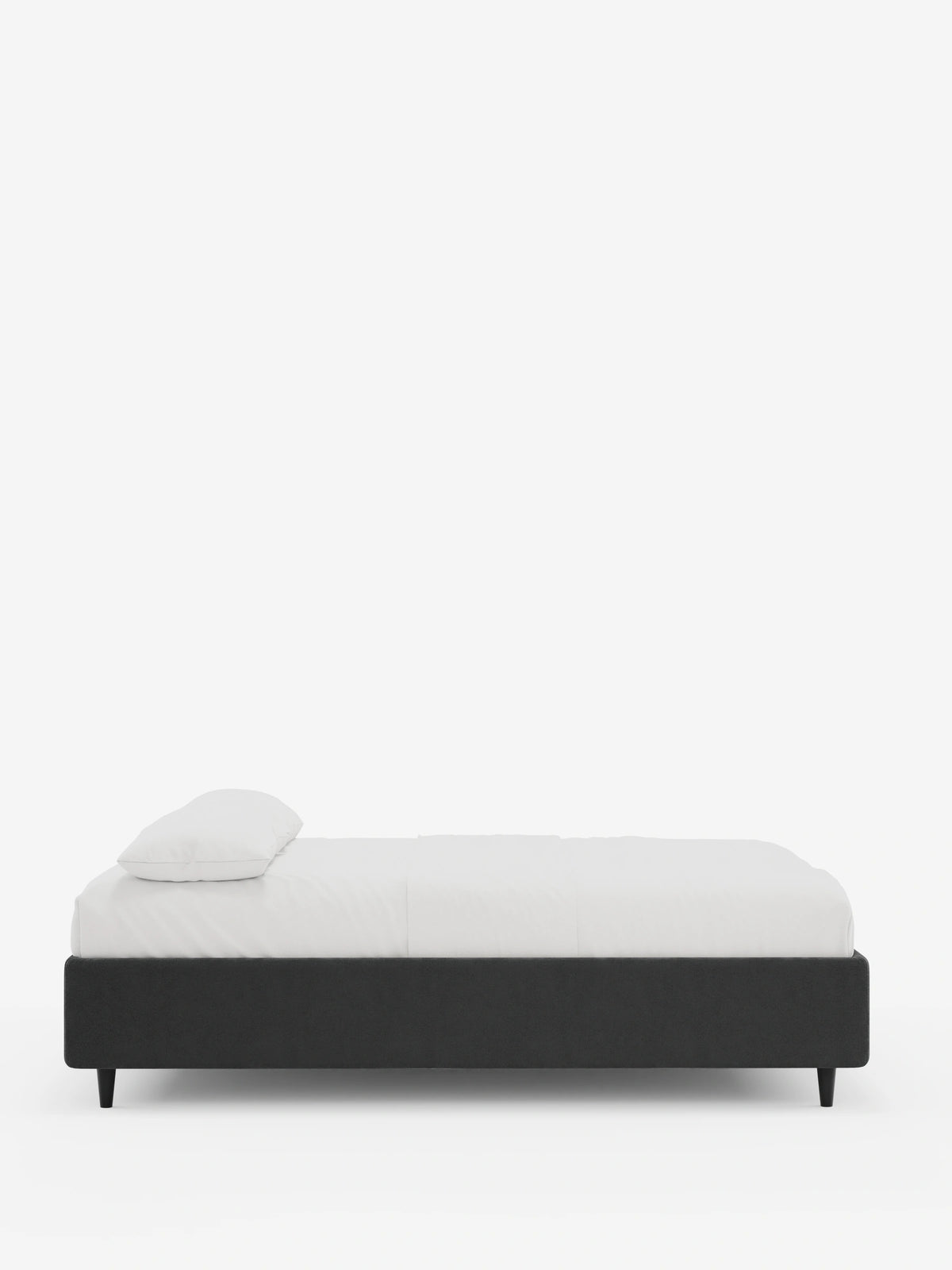 Vista laterale di un letto sommier imbottito ERON con vano contenitore in tessuto impermeabile certificato Oeko-Tex, con base imbottita color antracite scuro su quattro gambe cilindriche corte nere, su cui poggiano un materasso bianco e un cuscino singolo in uno studio.