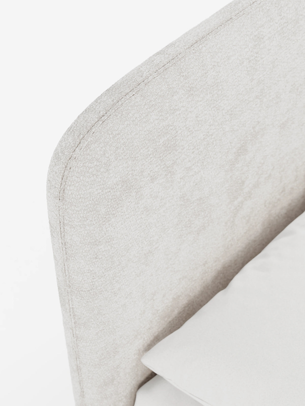 Un primo piano del letto imbottito KAELOS in stile minimal, caratterizzato da una testiera arrotondata e imbottita in tessuto beige chiaro con trama in rilievo e cuciture a vista lungo il bordo curvo, su uno sfondo bianco a tinta unita.