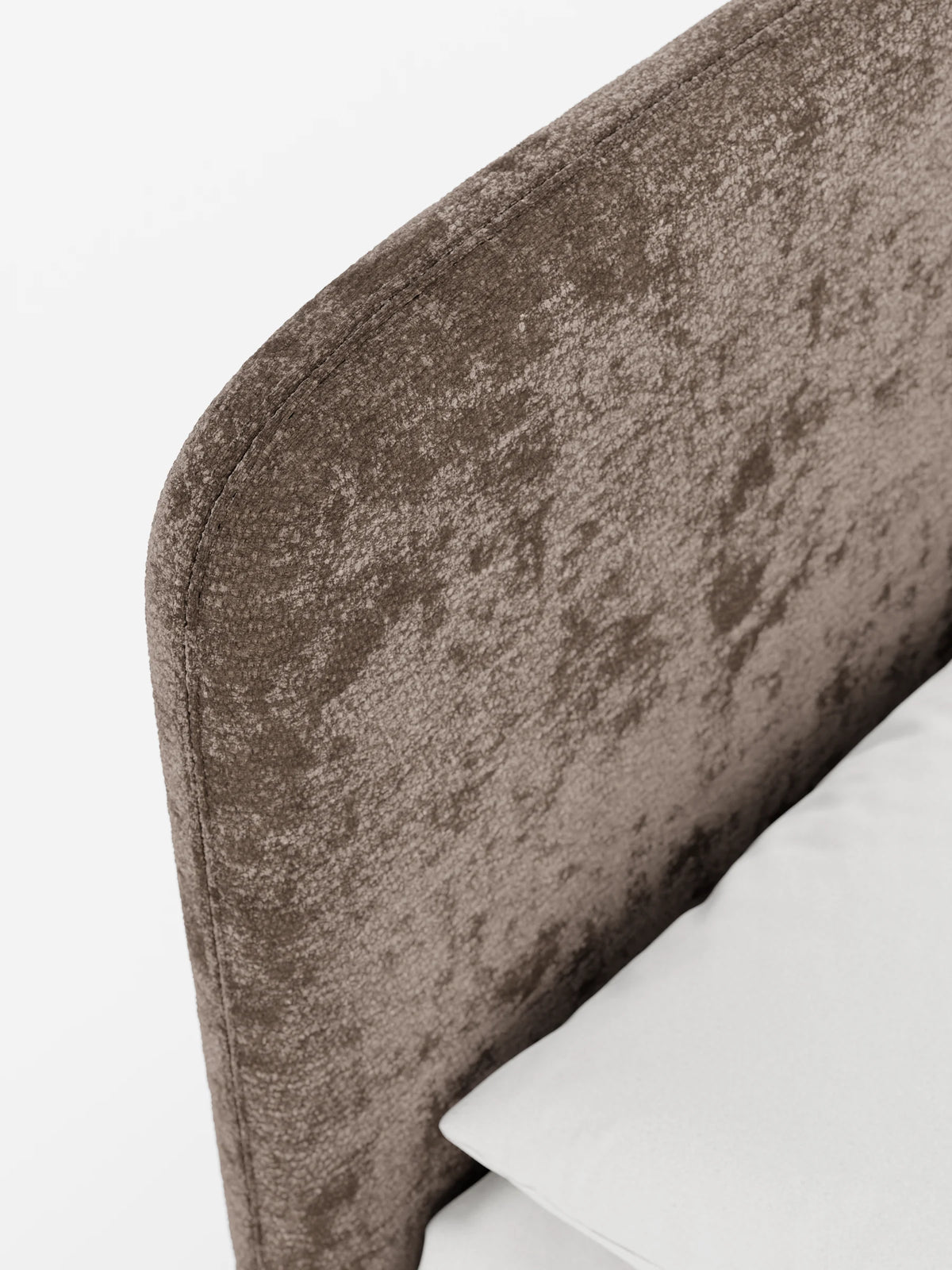 Un primo piano di un letto KAELOS minimal imbottito dalla forma arrotondata – con vano contenitore, gambe alte e testiera arrotondata in tessuto ciniglia – caratterizzato da un tessuto ciniglia marrone a trama rigata, una superficie imbottita curva e un angolo con cuscino bianco su sfondo bianco.