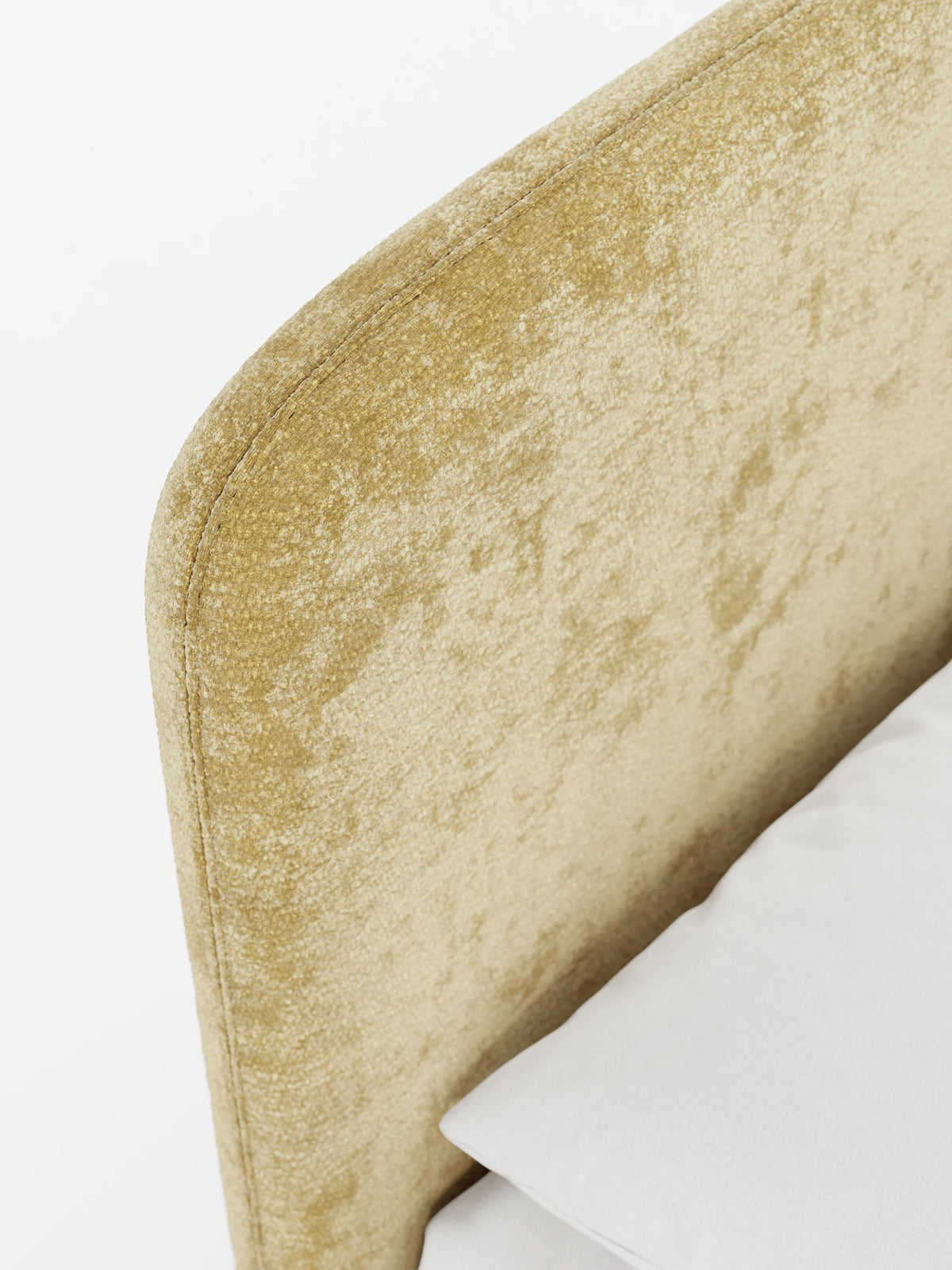 Un primo piano di un letto KAELOS minimal imbottito con una testiera arrotondata in ciniglia giallo-dorata e un cuscino bianco a tinta unita, su uno sfondo bianco uniforme.