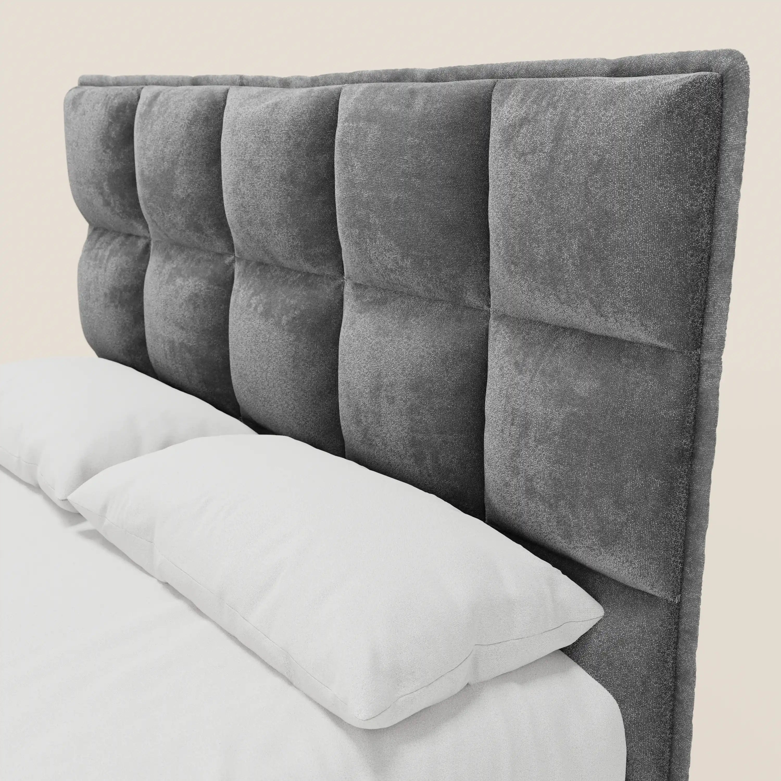 Il letto imbottito AURO di Letti.store presenta una testiera alta e capitonné in ciniglia grigia certificata Oeko-Tex® con motivo a griglia. Due cuscini bianchi e la biancheria da letto ne esaltano l'elegante trama, che risalta sullo sfondo a tinta unita.