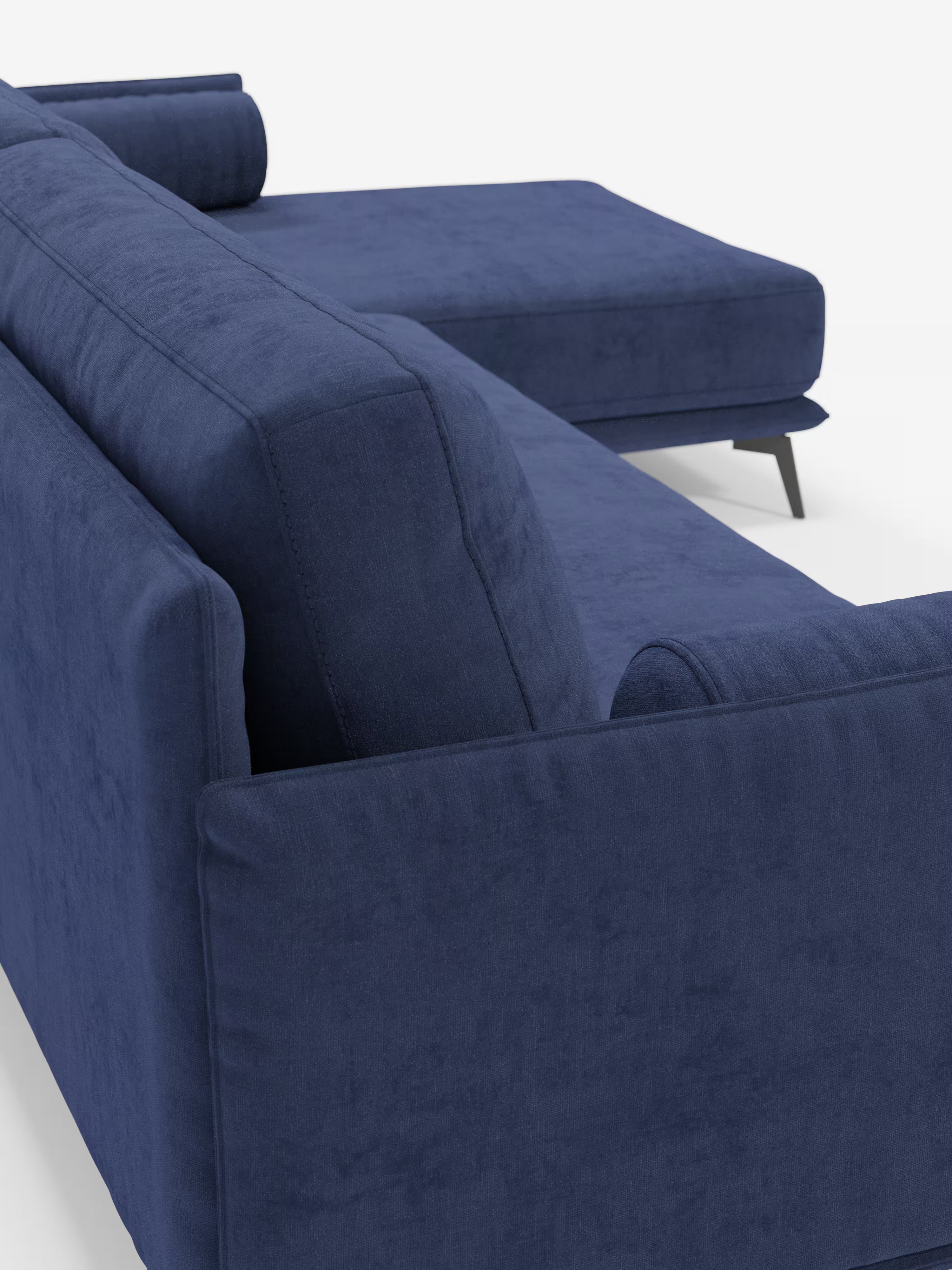 Primo piano del divano moderno angolare con penisola “Basalto” di Divani.Store, rivestito in tessuto T31 blu scuro e resistente alle macchie. Questo elegante divano componibile presenta ampi braccioli rettangolari, morbidi cuscini in piuma e una robusta base in metallo con sottili gambe nere inclinate. La chaise longue si estende verso destra e due cuscini cilindrici completano il design raffinato di questo divano contemporaneo.